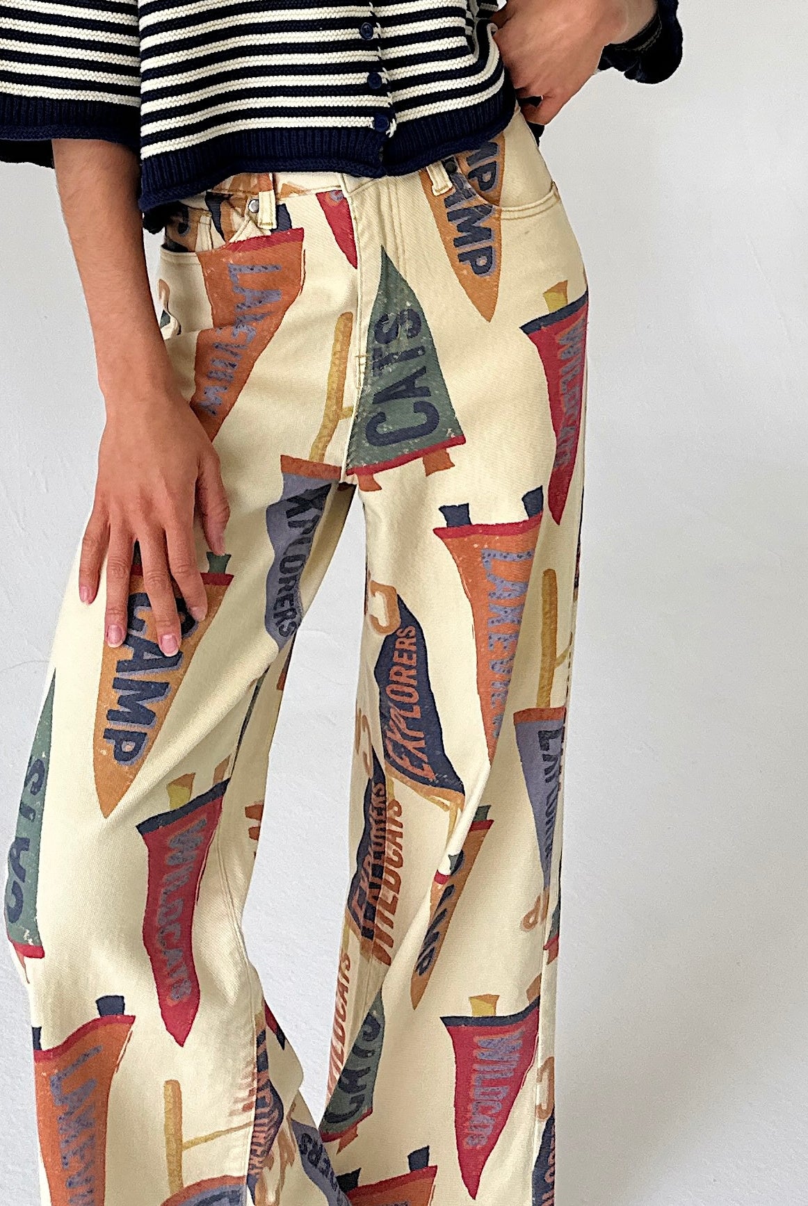 A person wearing wide-leg cream pants with an allover vintage pennant flag print in red, blue, orange, and brown, paired with a black-and-white striped top against a plain light background.