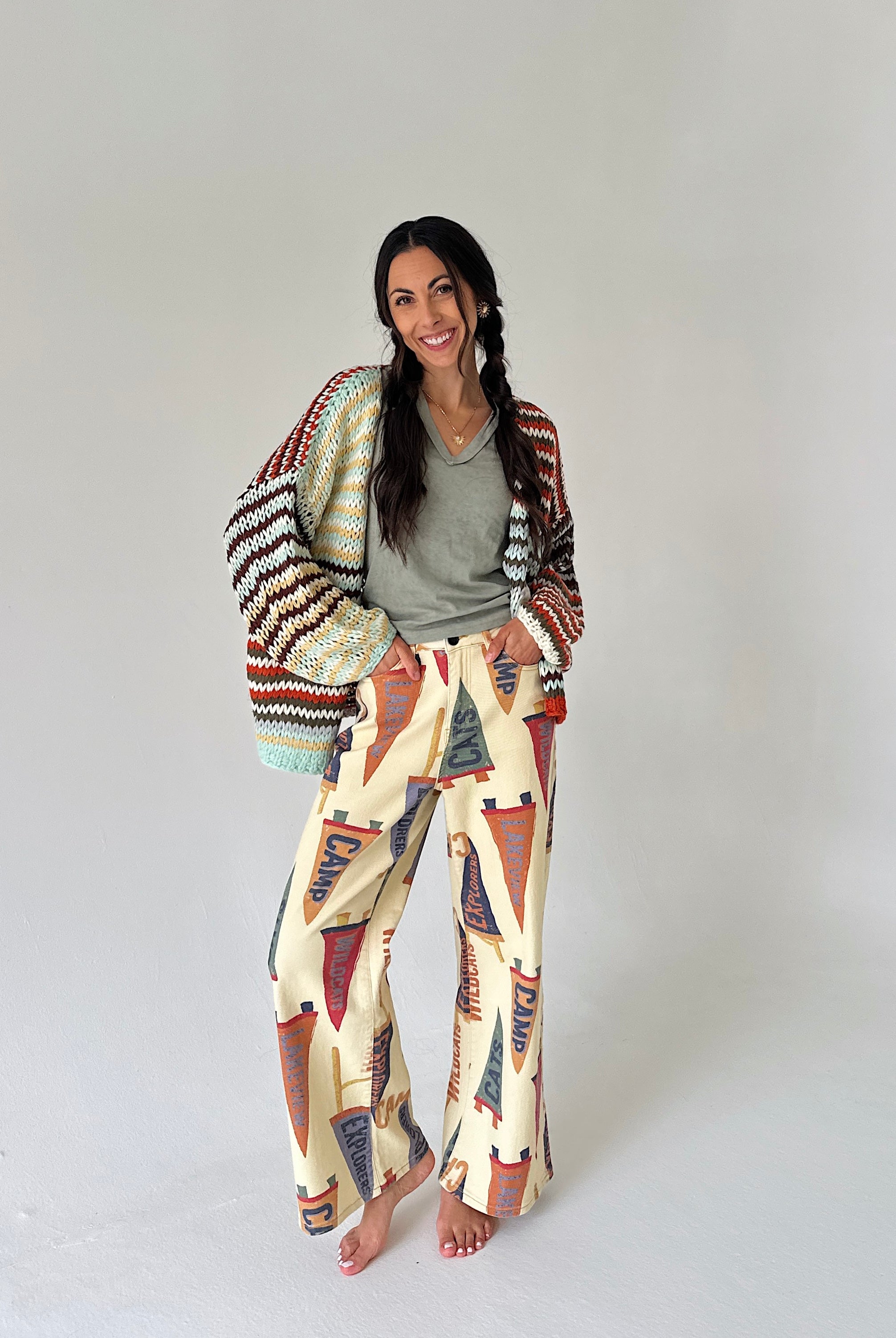 A woman stands barefoot against a plain light gray studio background, wearing cream wide-leg pants with a colorful pennant print, a green top, and a multicolor knit cardigan.