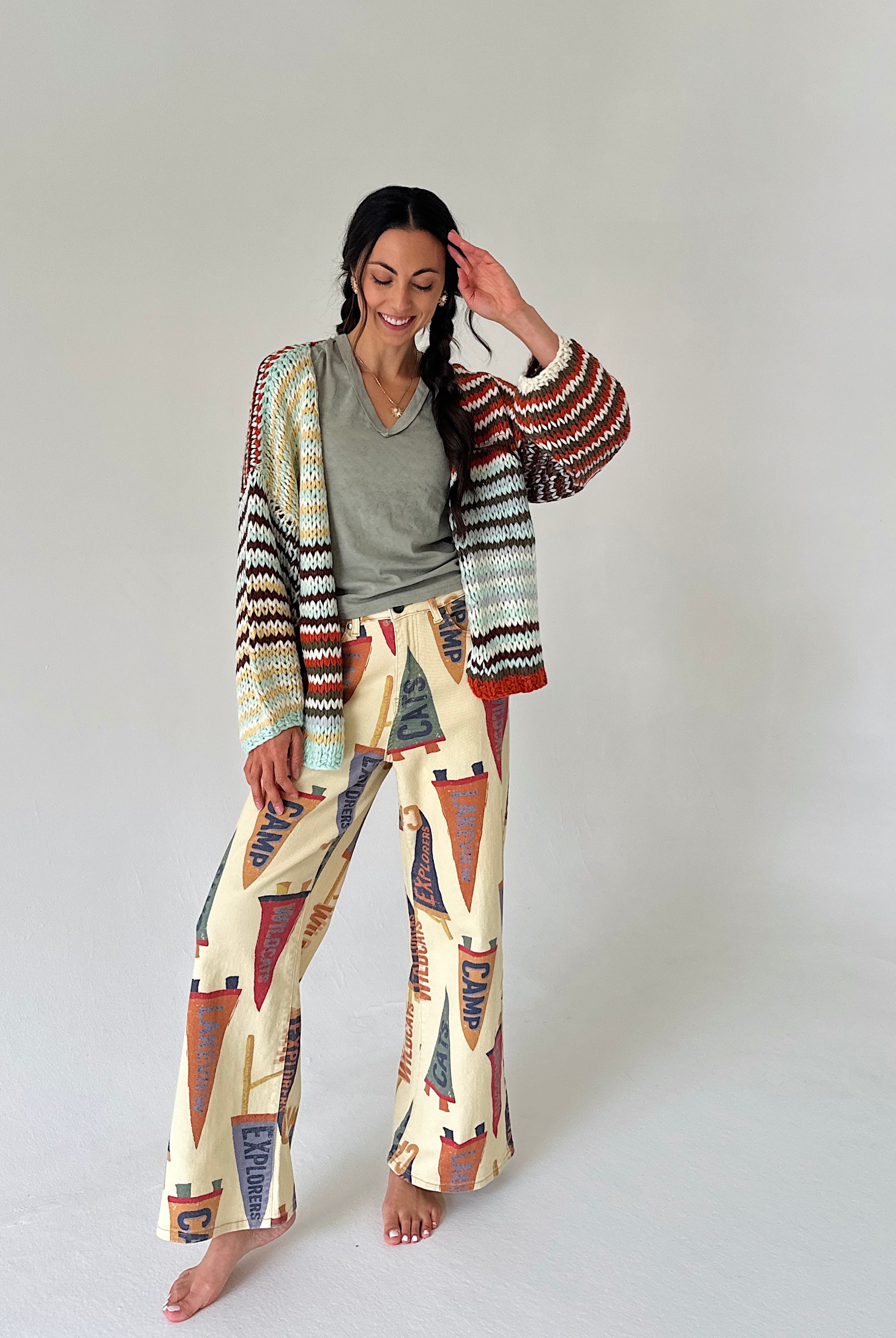 A woman stands barefoot against a plain light gray studio background, wearing a colorful striped knit cardigan and wide-leg pants set with a cream base and pennant multicolor patterns in rust, navy, olive, and black.