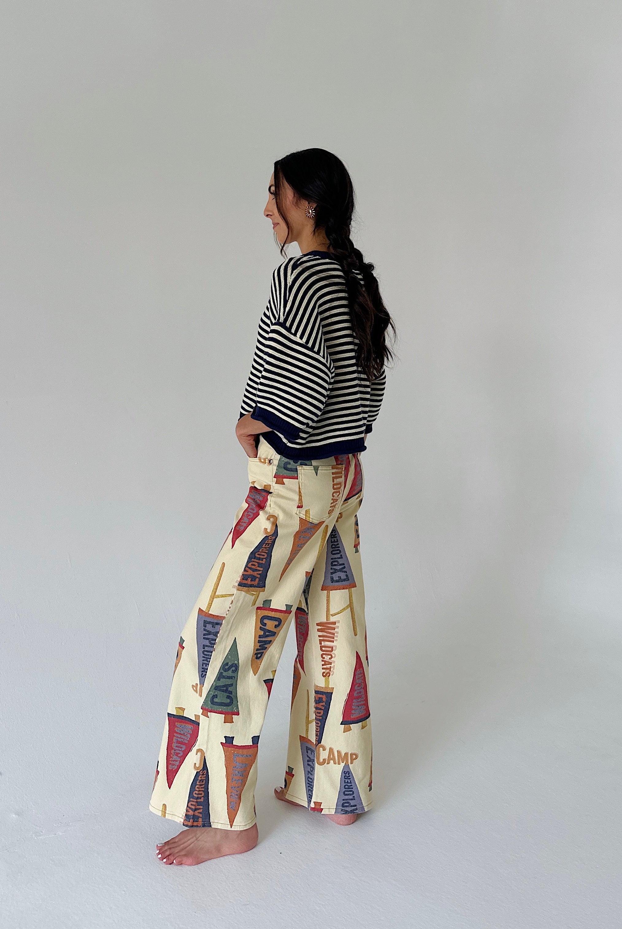 A woman stands barefoot in profile wearing a black and white striped cardigan and cream wide-leg pants with a colorful vintage flag print, set against a plain light gray studio background.