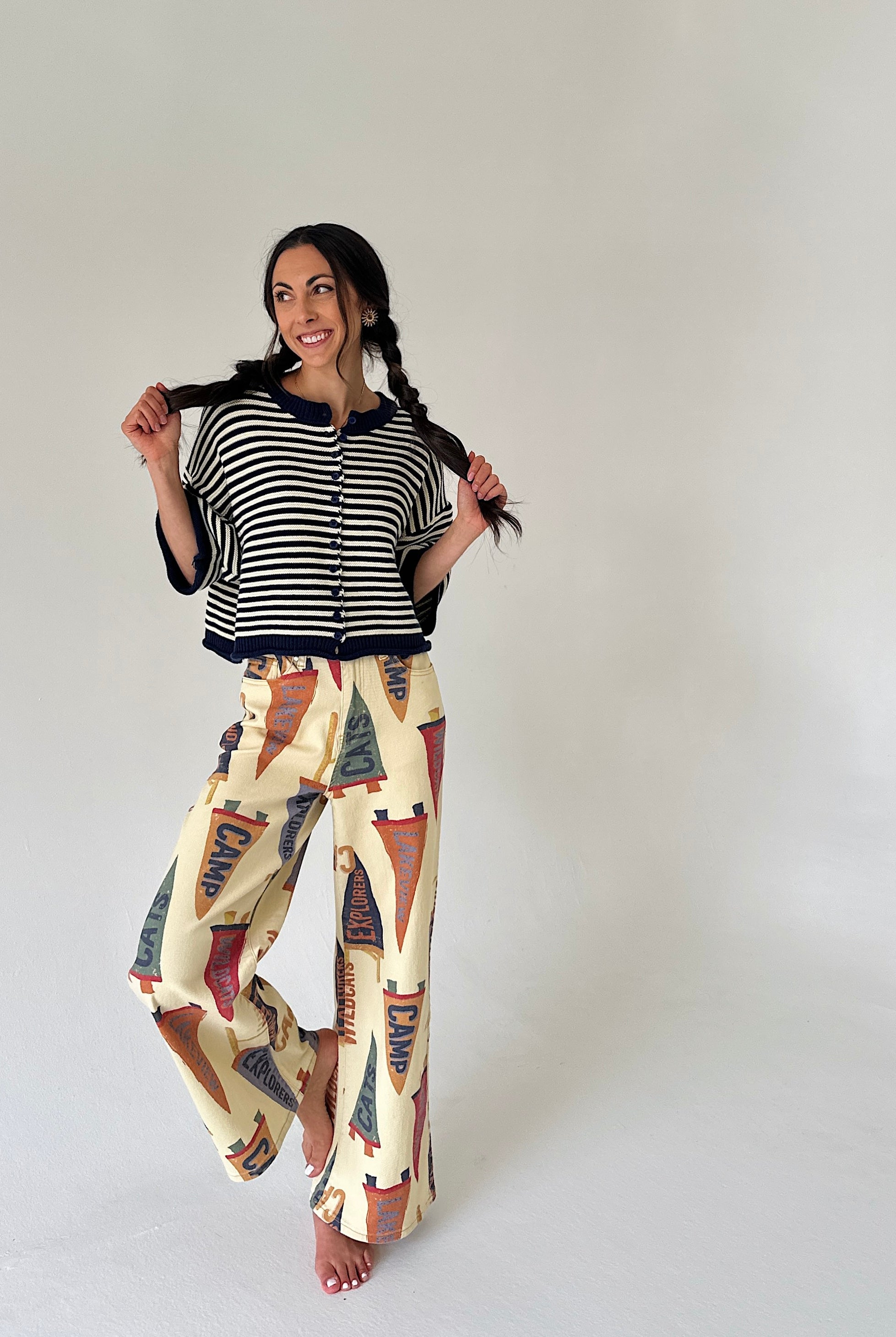 A woman modeling a two-piece outfit consisting of a navy and white striped short-sleeve cardigan with a button-up front and cream wide-leg pants featuring a colorful pennant flag pattern, set against a plain light gray studio background.