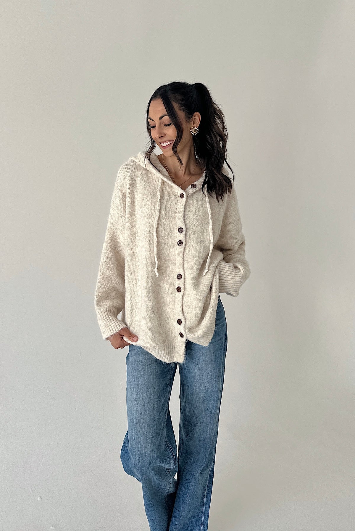 A woman wearing a cream-colored, fuzzy button-up hooded sweater and blue jeans, standing against a plain light gray studio background.