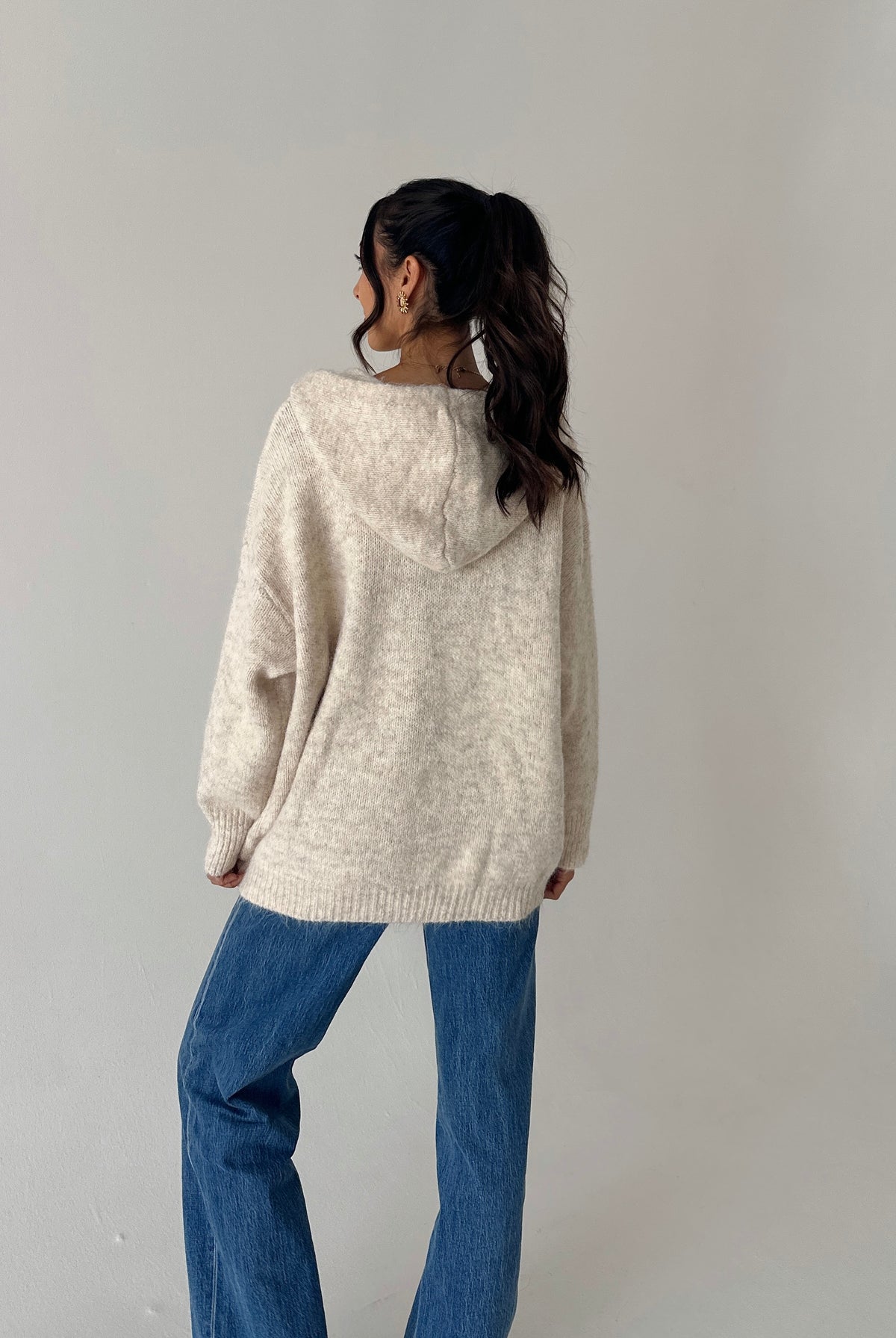 A woman shown from the back wearing a cream-colored, oversized knit hoodie sweater and blue jeans, standing against a plain light gray wall.