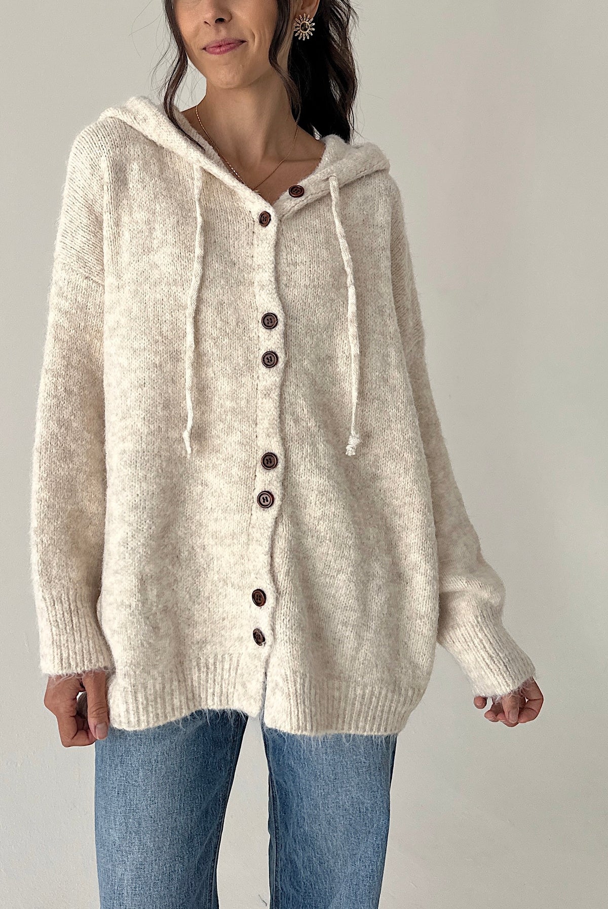 A woman wearing a cream-colored, fuzzy knit hooded button-up sweater with brown buttons and matching drawstrings, paired with blue jeans, against a plain light gray background.