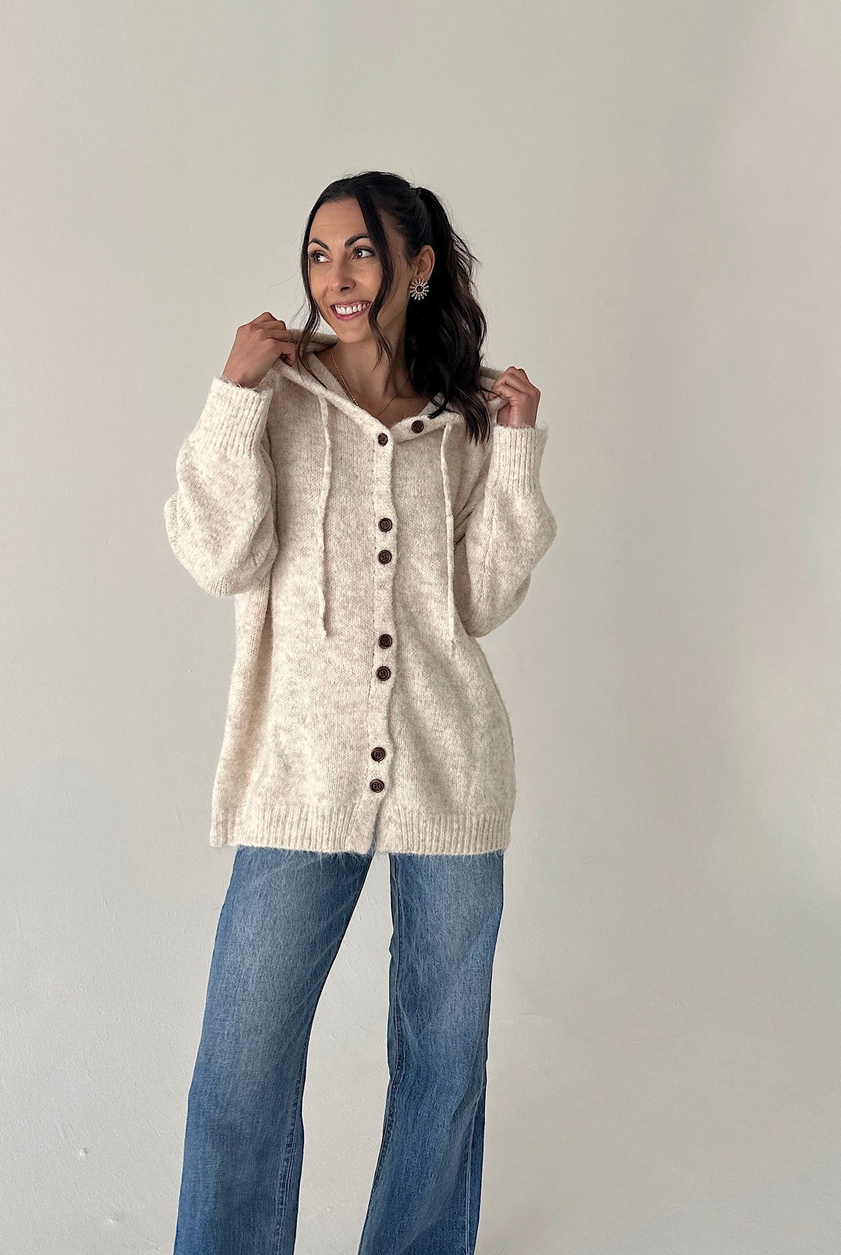 A woman modeling a cream-colored, oversized knit cardigan with a hood and front buttons, paired with blue jeans, against a plain light gray studio background.