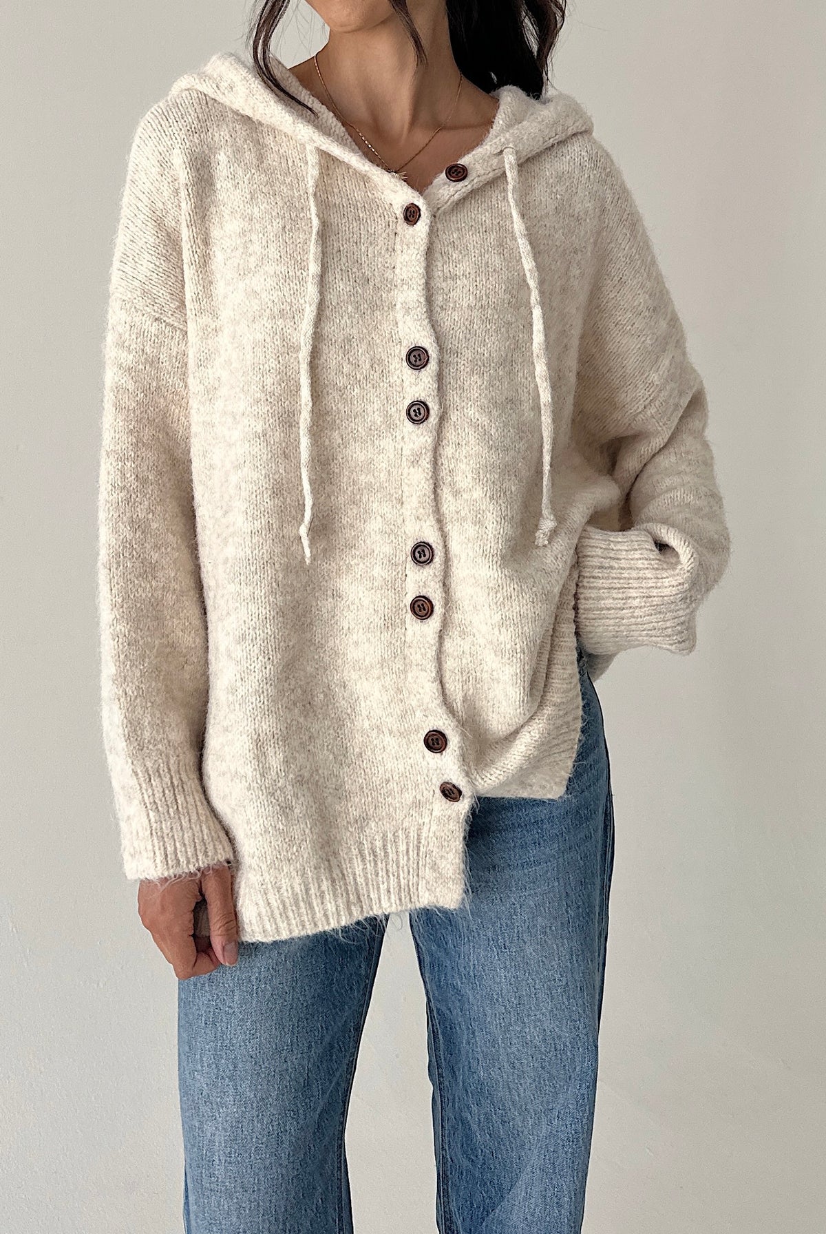 A person wearing a chunky knit hooded cardigan in a warm off-white/cream color with dark brown buttons, paired with blue jeans, against a plain light gray background.
