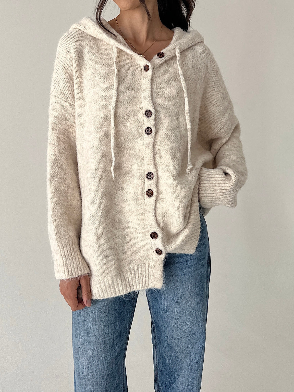 A person wearing a chunky knit hooded cardigan in a warm off-white/cream color with dark brown buttons, paired with blue jeans, against a plain light gray background.