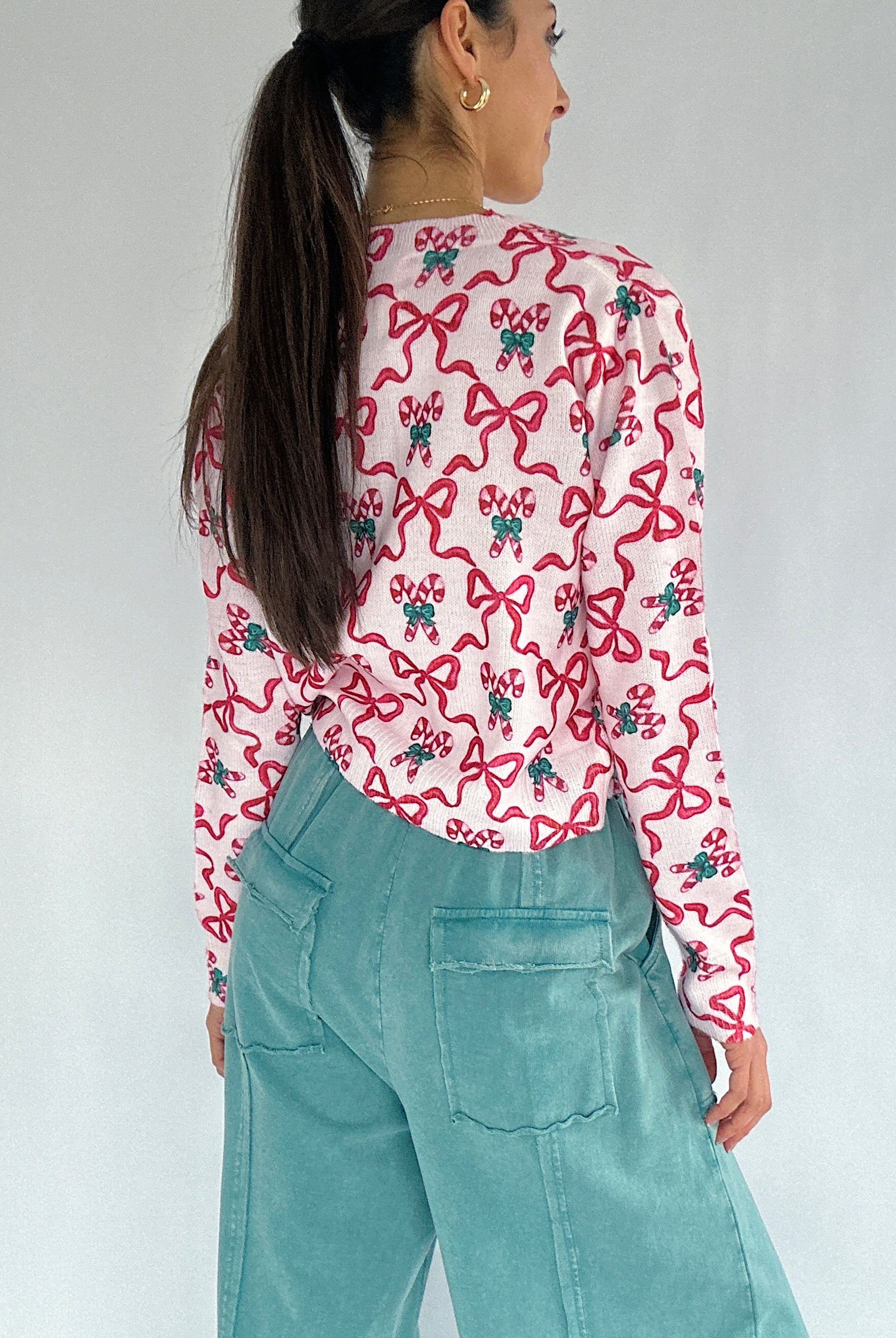 Person wearing a cardigan with red bow and candy cane pattern and teal pants against a plain background