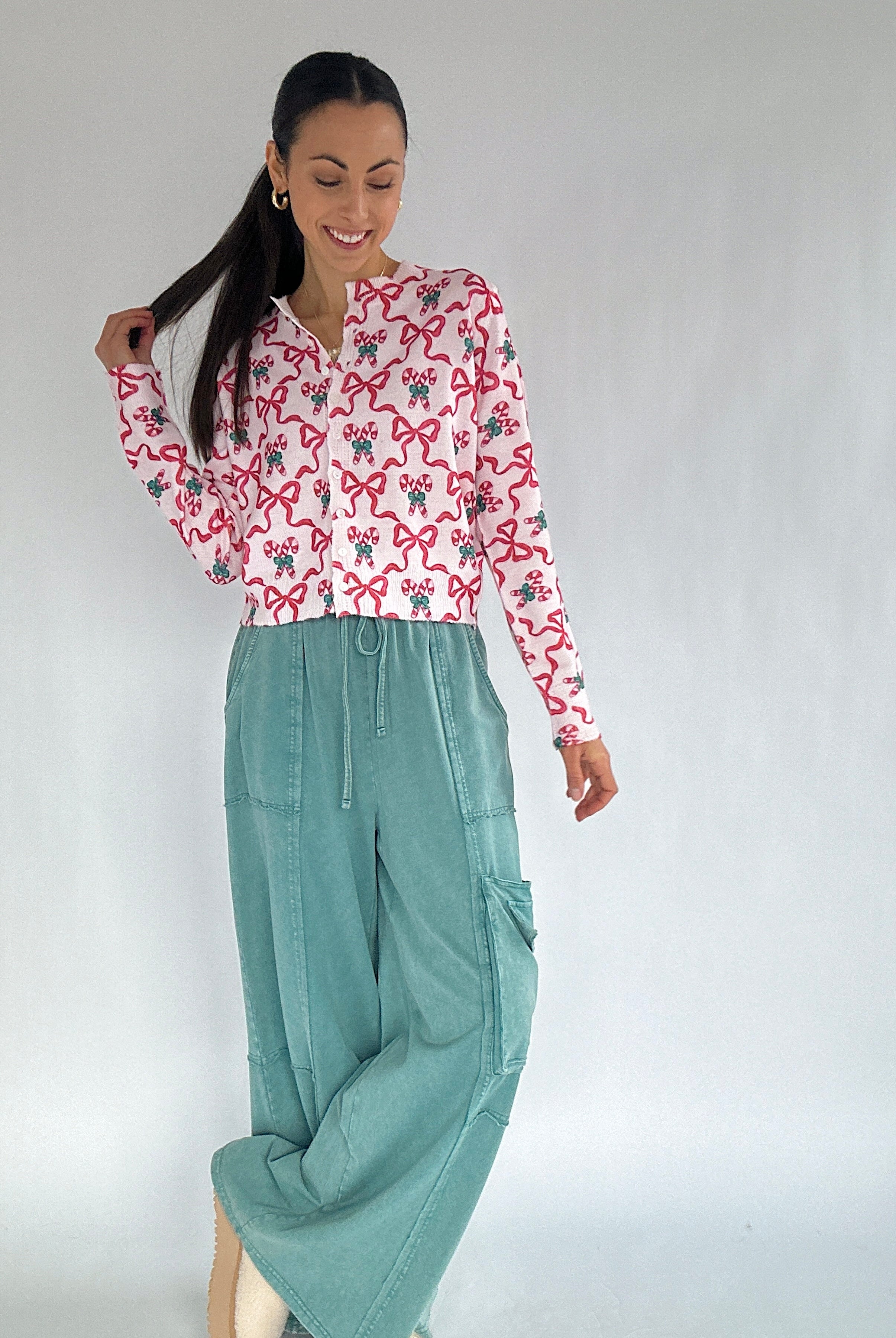 Woman wearing a candy cane and bow patterned cardigan top and teal pants against a plain background