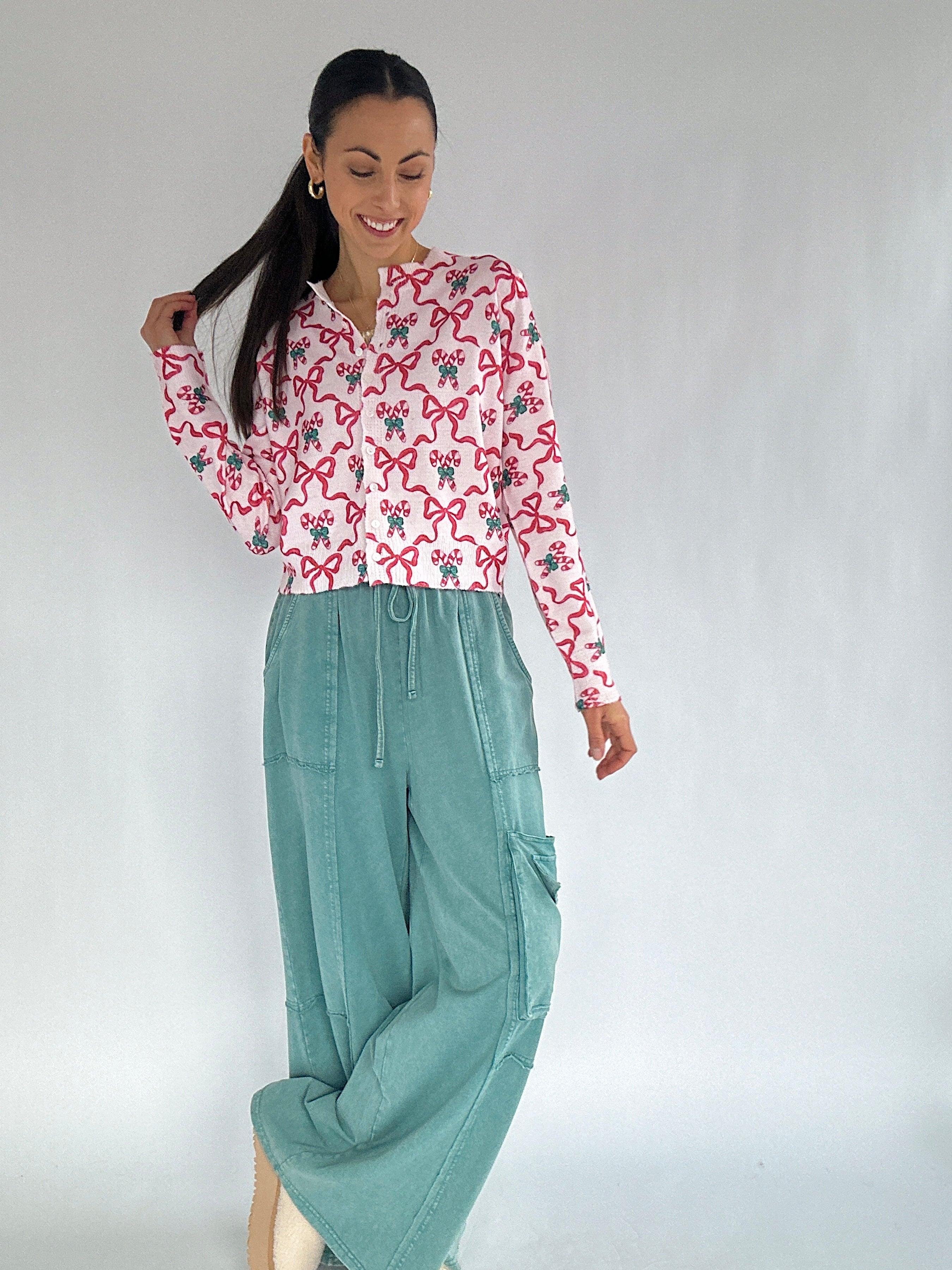Woman wearing a candy cane and bow patterned cardigan top and teal pants against a plain background