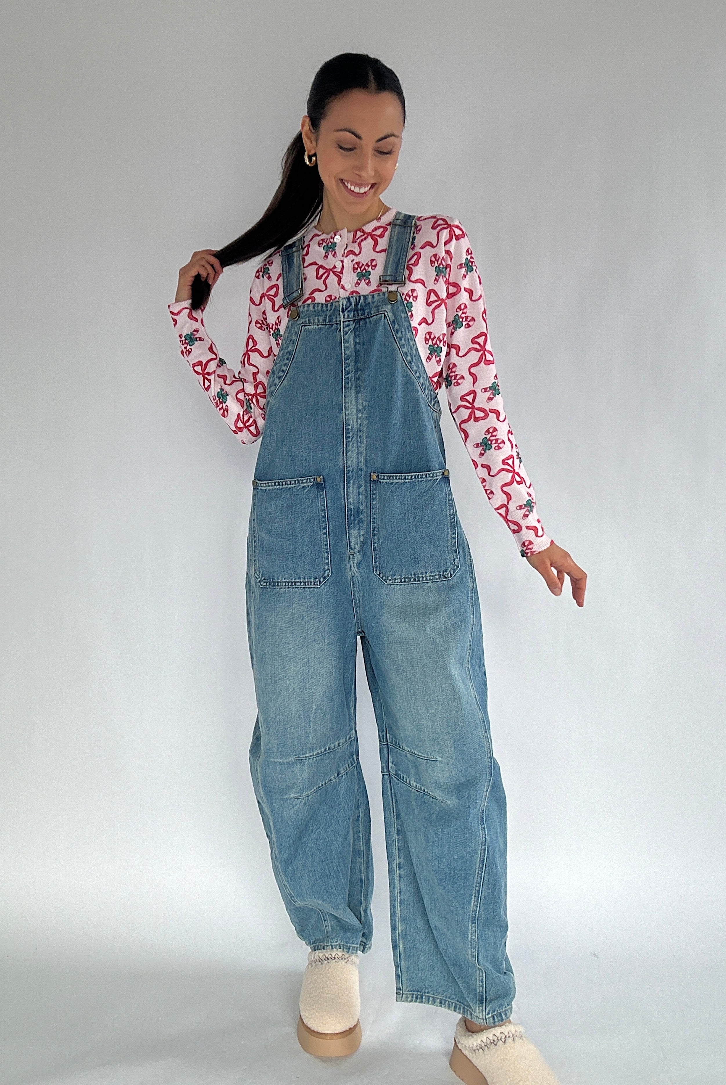 Woman wearing a candy cane and bow long-sleeve cardigan under denim overalls on a plain background