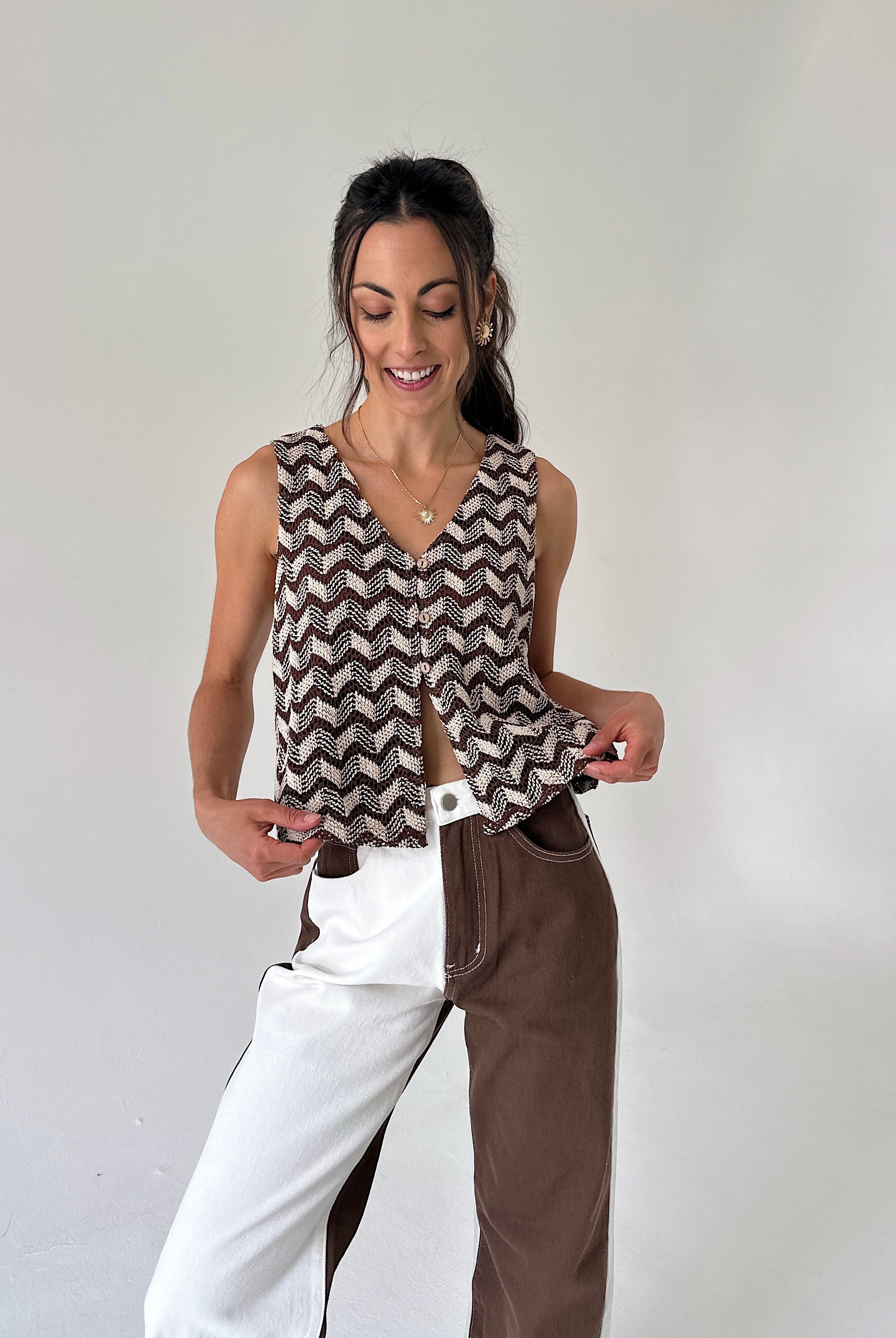 A woman modeling a sleeveless V-neck top with a brown and beige chevron pattern, paired with a split-color pants outfit, against a plain light gray background.