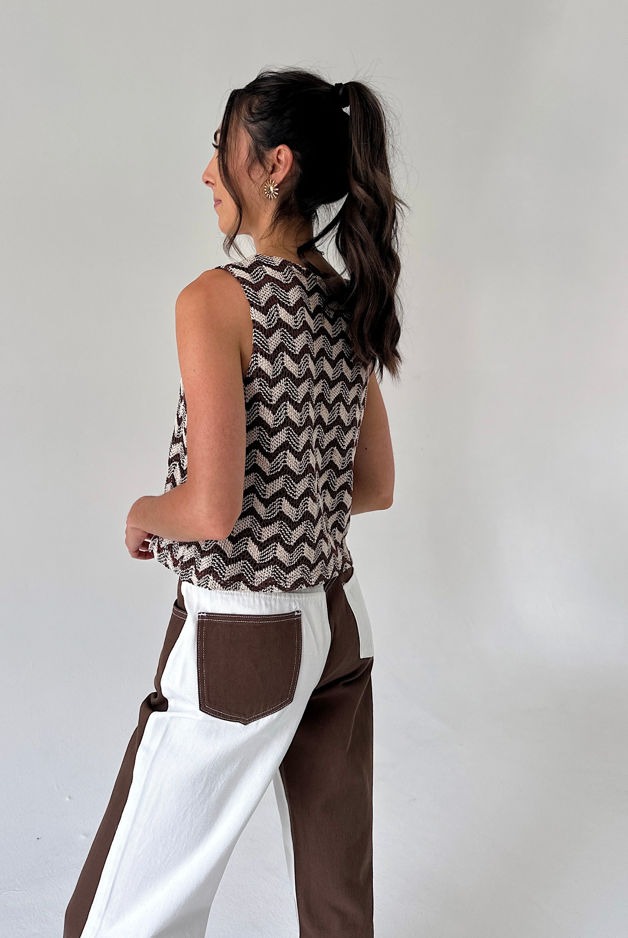 A woman shown from the back wearing a sleeveless knit top with a brown and white chevron pattern, paired with white and brown color-blocked pants, against a plain light gray background.