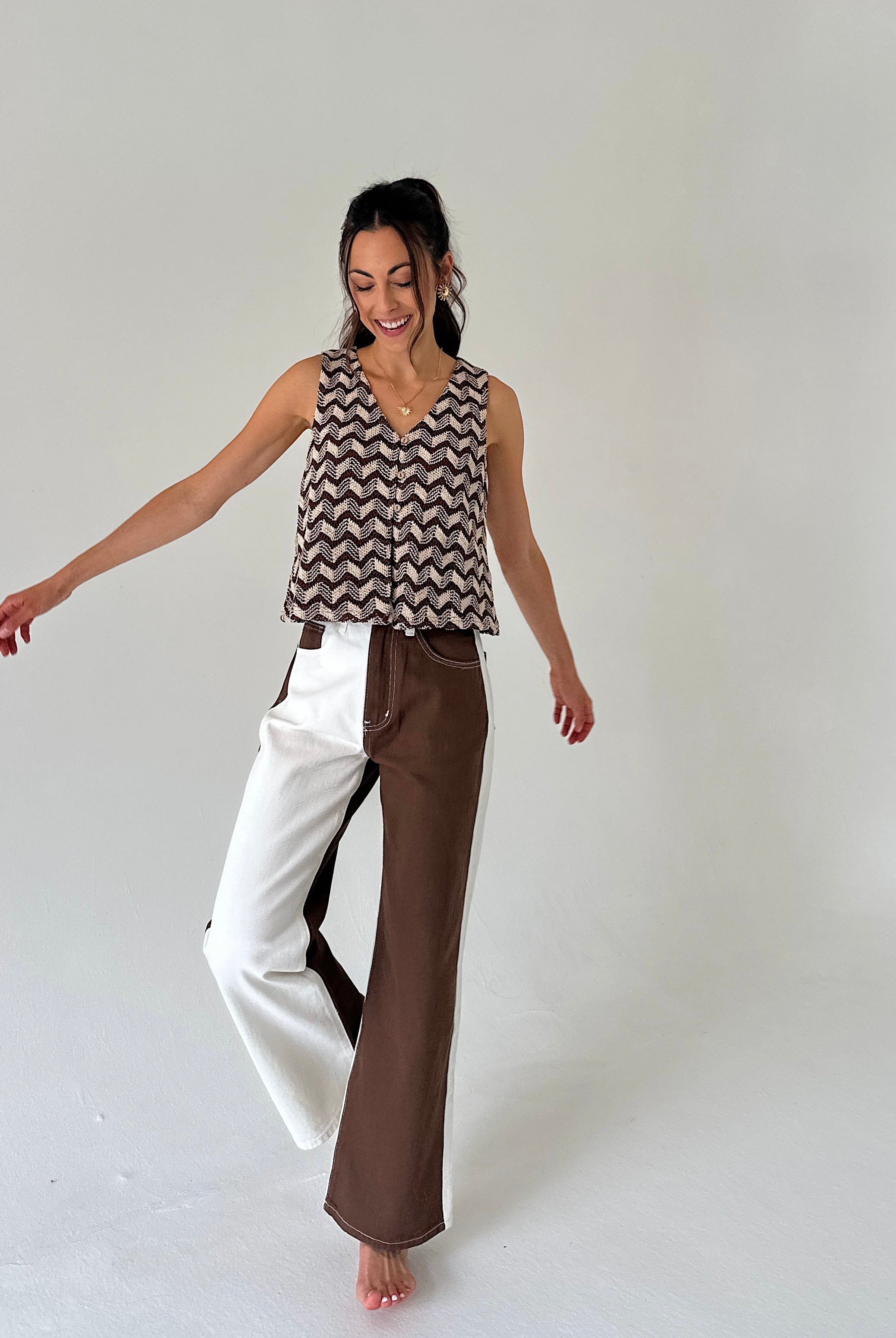 A woman modeling a sleeveless V-neck top with a brown and beige chevron pattern, paired with wide-leg pants split down the middle into white and brown sections, against a plain light gray studio background.