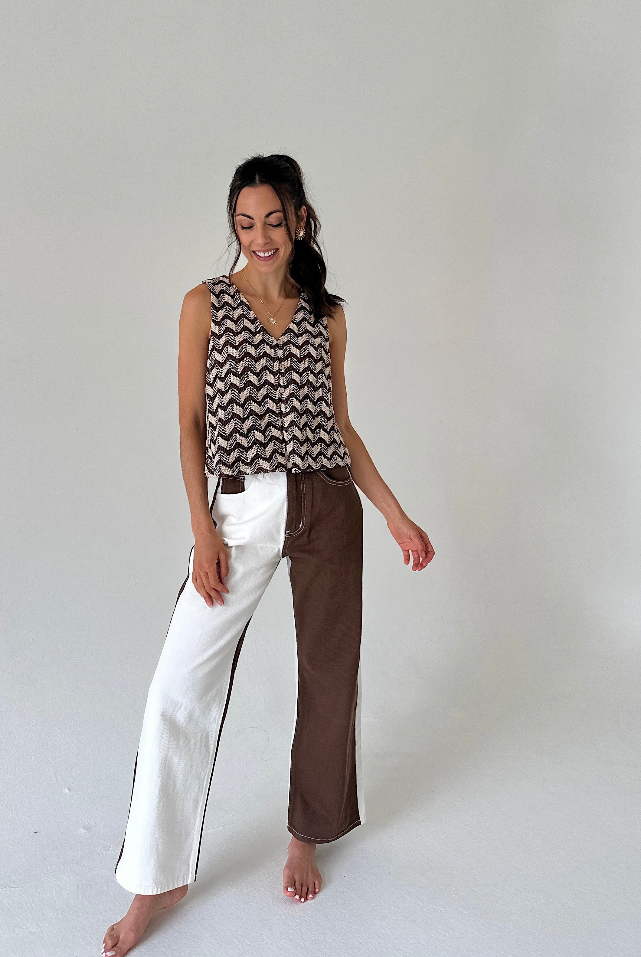 A woman stands barefoot against a plain light gray studio background, wearing a sleeveless top with a brown and beige chevron pattern and wide-leg pants split down the middle with one leg in white and the other in brown.