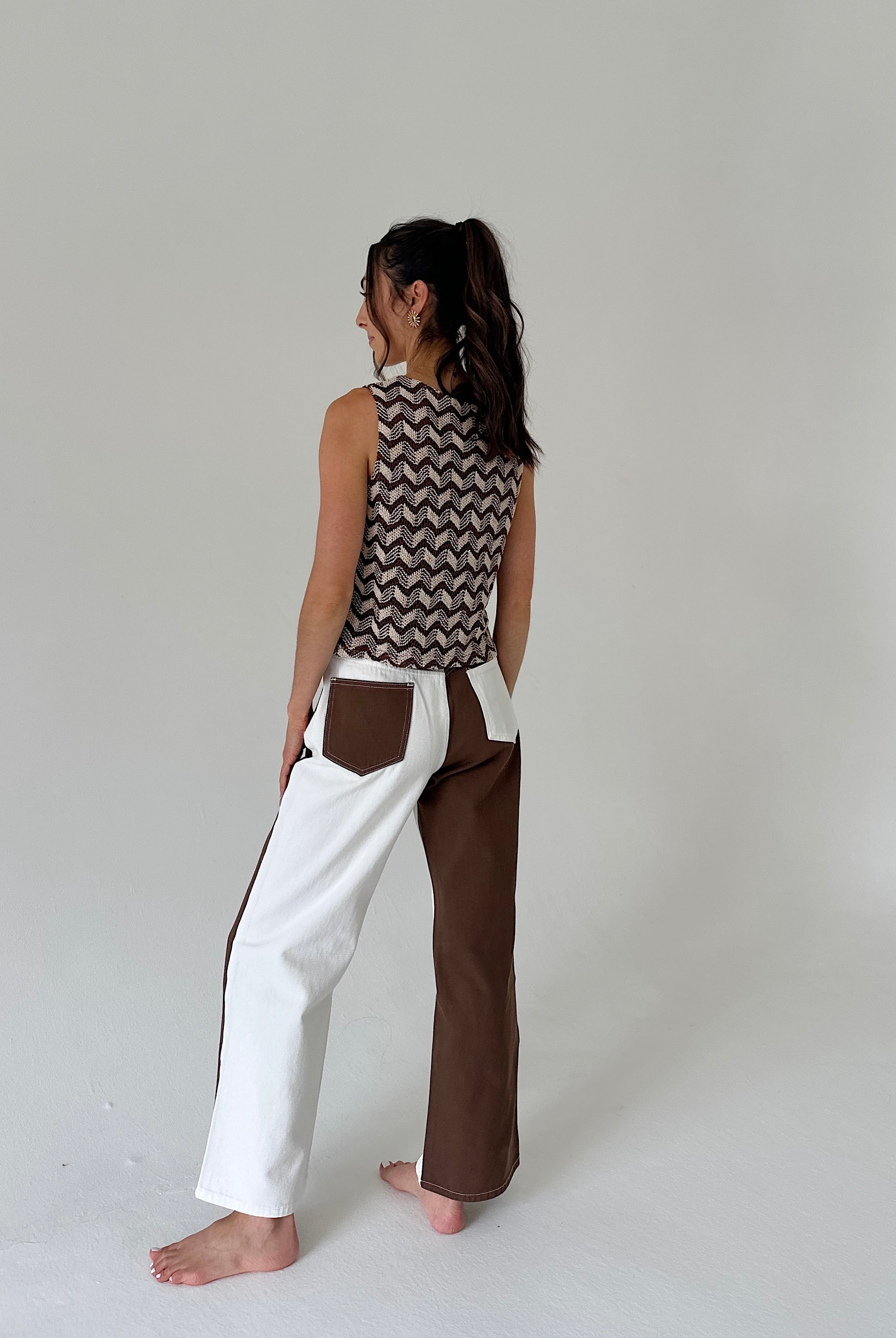 A woman stands barefoot in a studio wearing a sleeveless top with a brown and beige chevron pattern and wide-leg pants with a color-blocked design in white and brown.