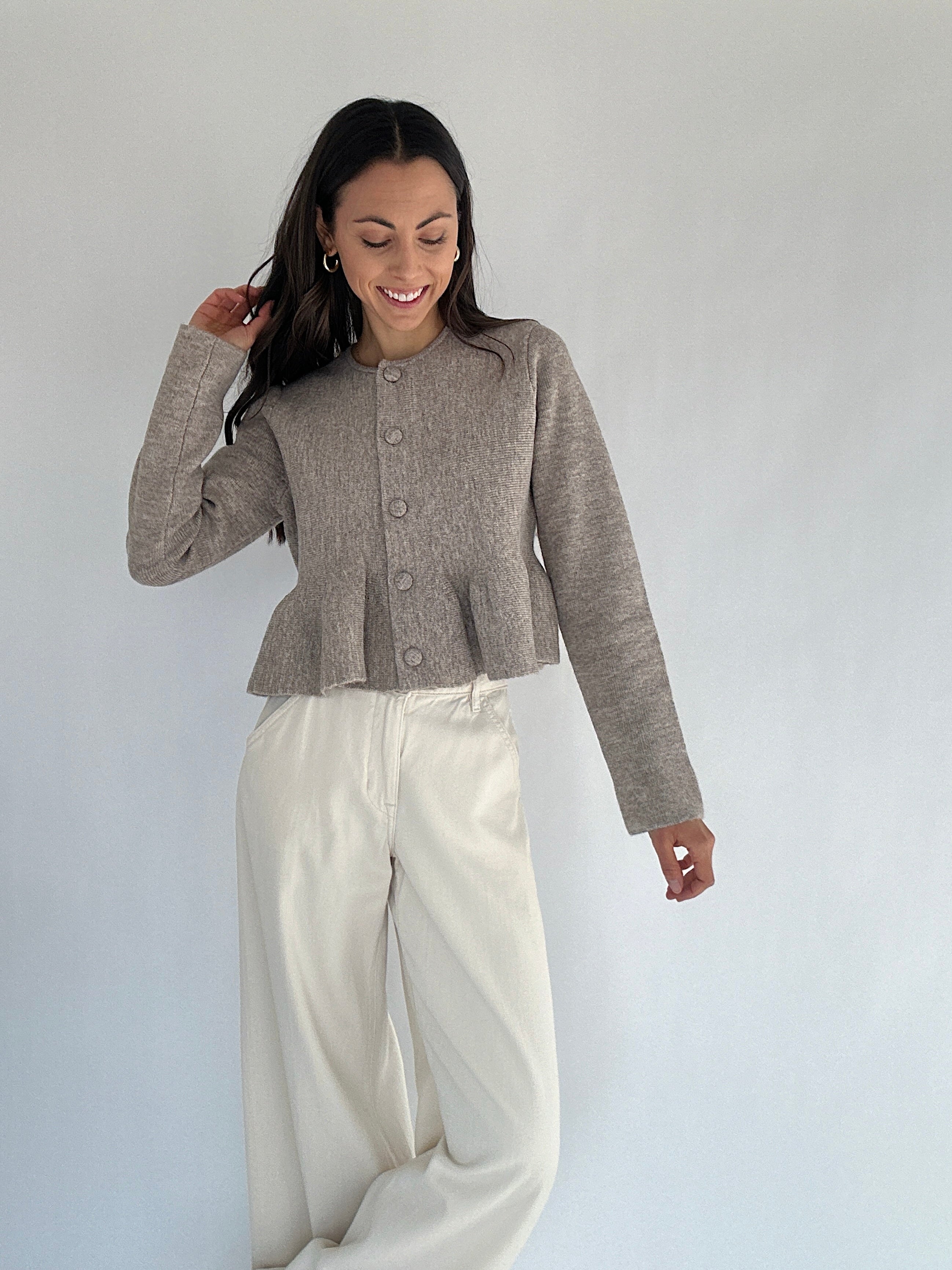 Woman wearing a brown peplum button front cardigan and white pants against a plain background