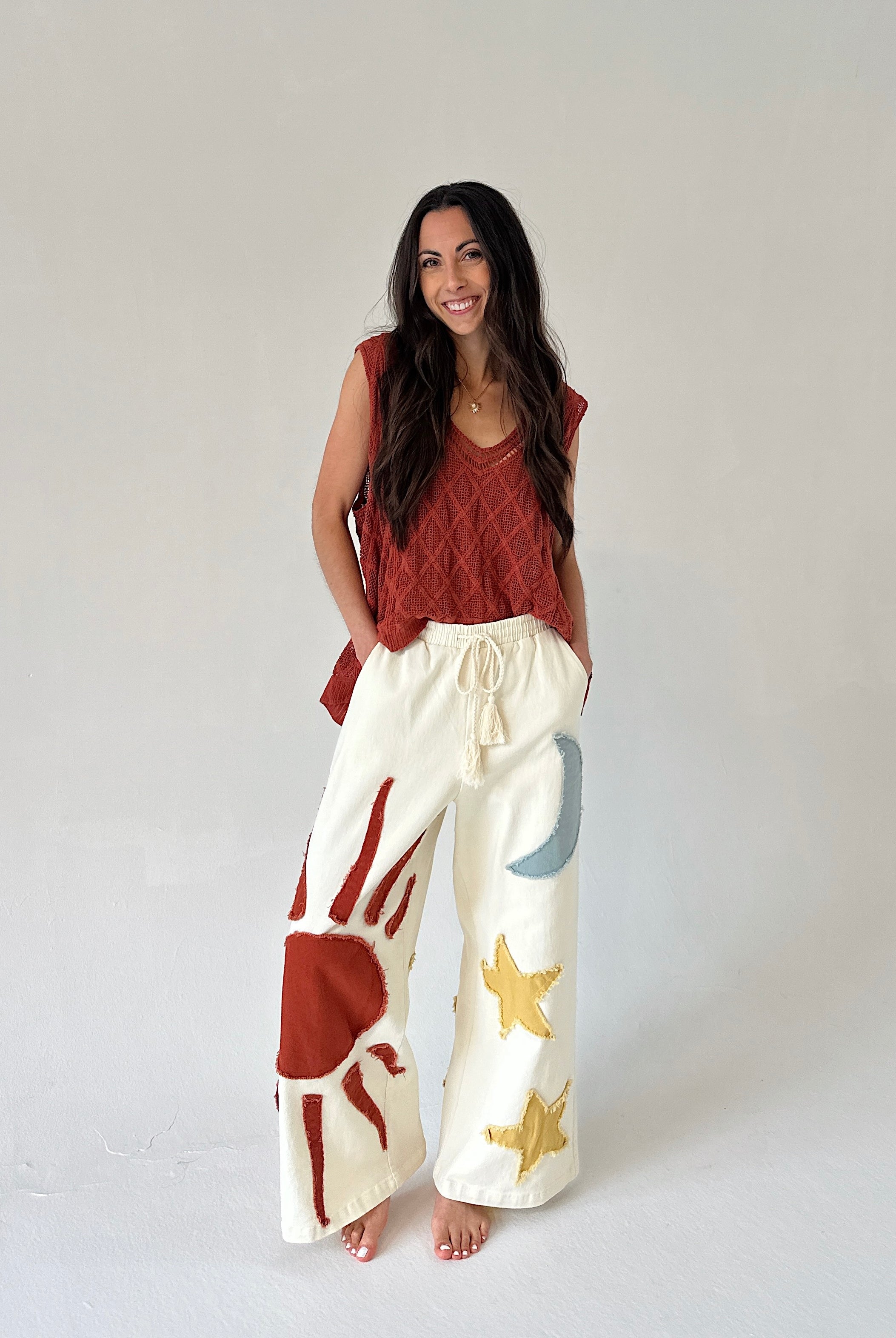 Woman wearing a rust-colored top and white pants with sun, moon and star designs on a plain background