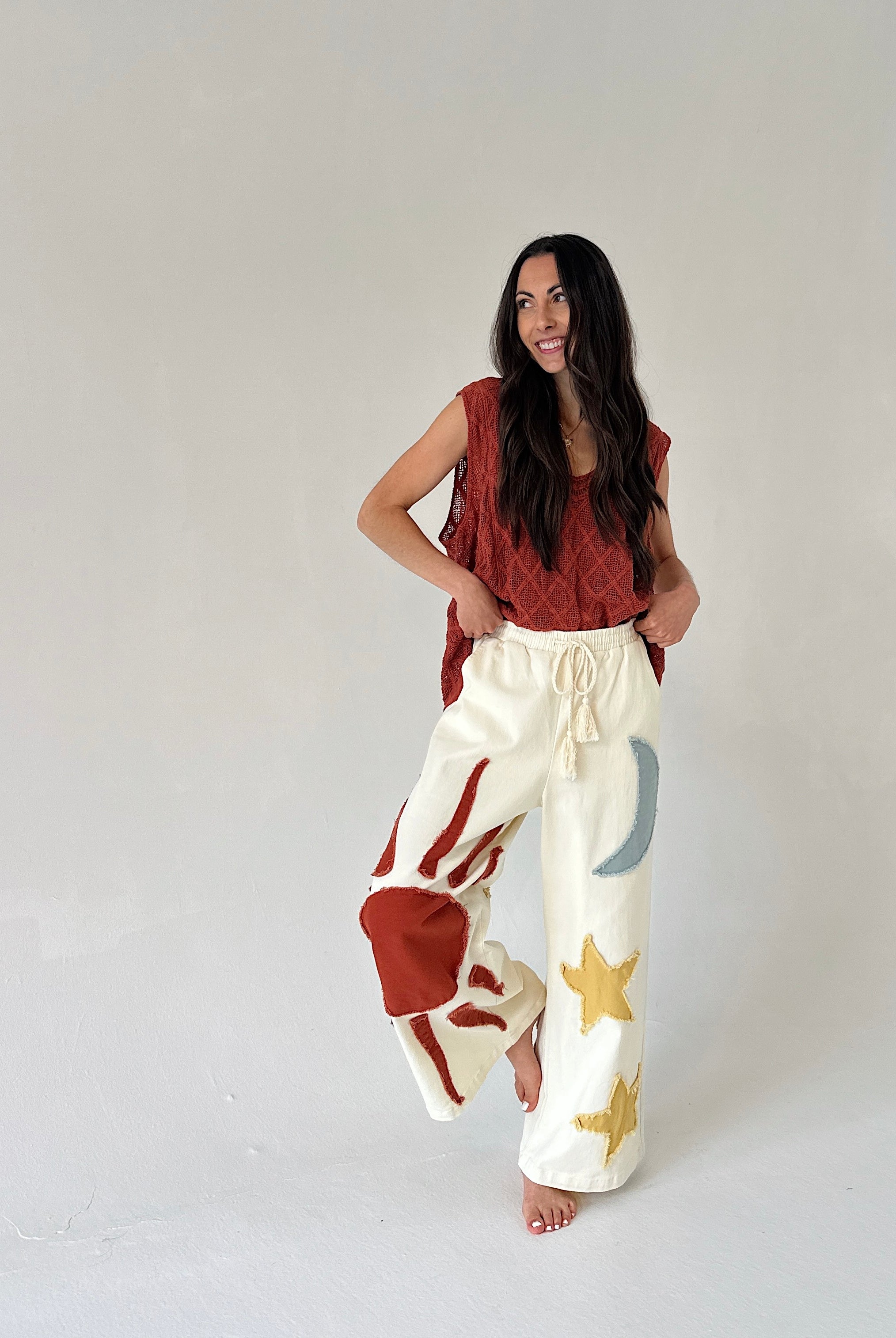 Woman wearing a rust-colored sleeveless top and white pants with star patterns on a plain background