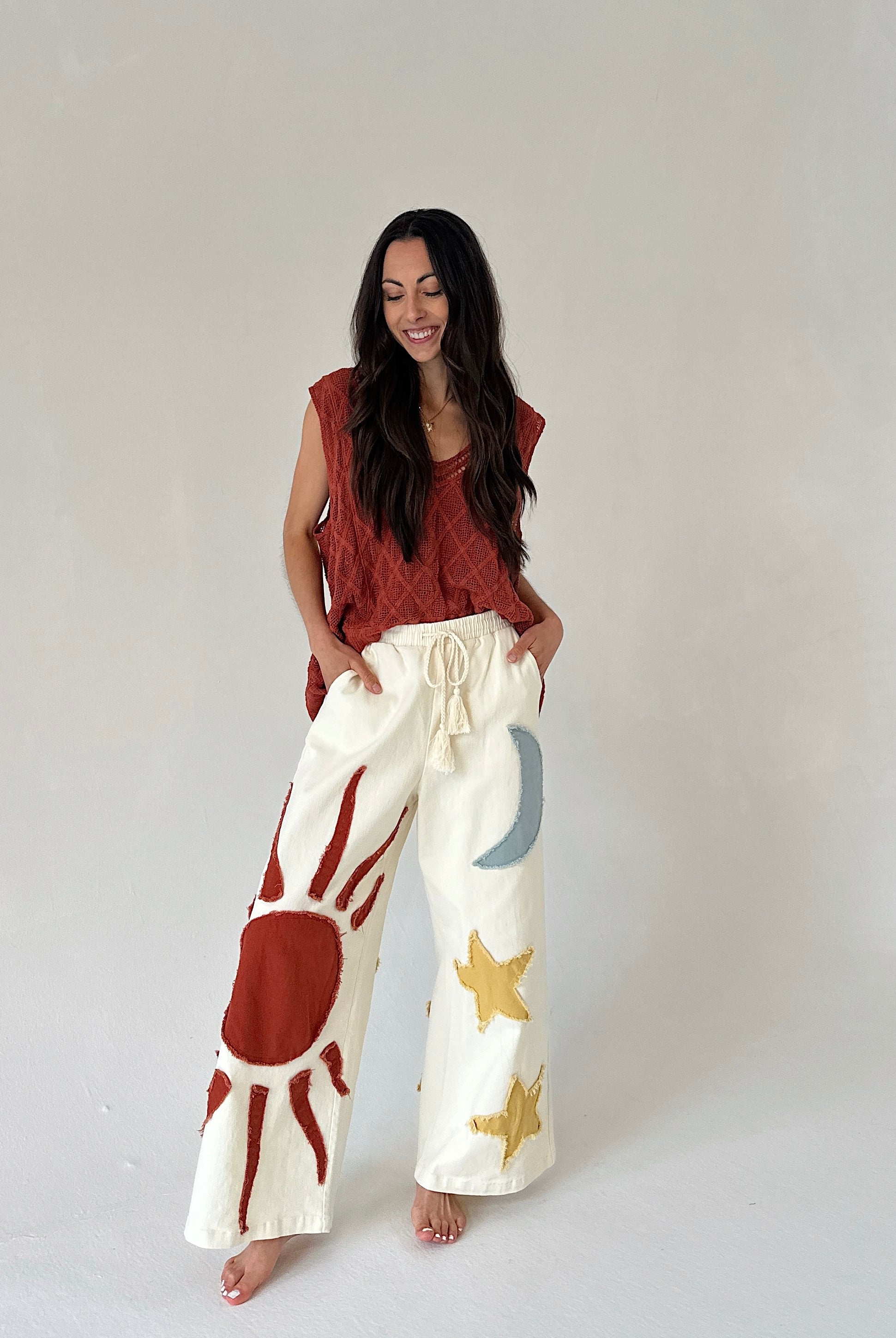 Woman wearing a rust-colored sleeveless top and white pants with sun and star patterns on a plain background