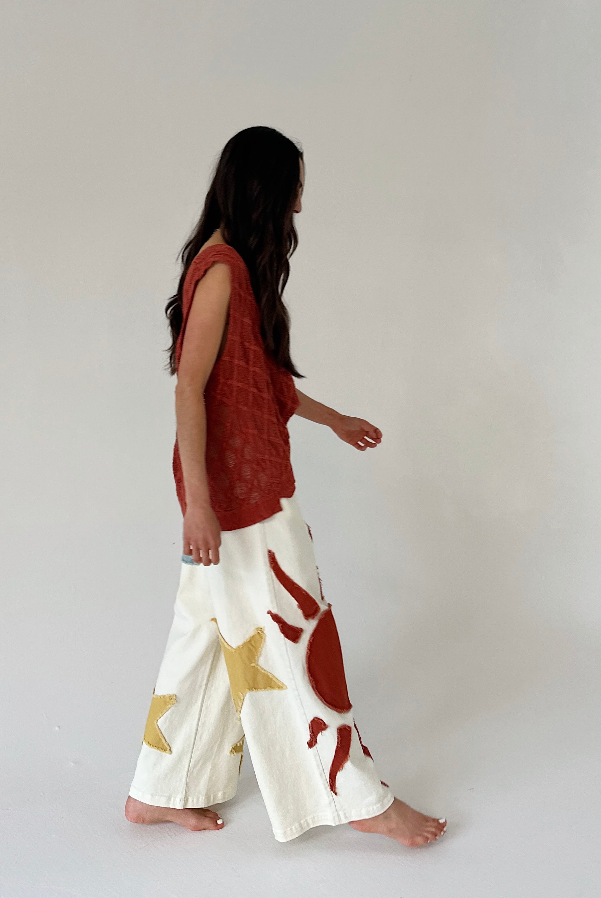 Woman wearing a rust-colored top and white pants with red and gold patterns on a plain background