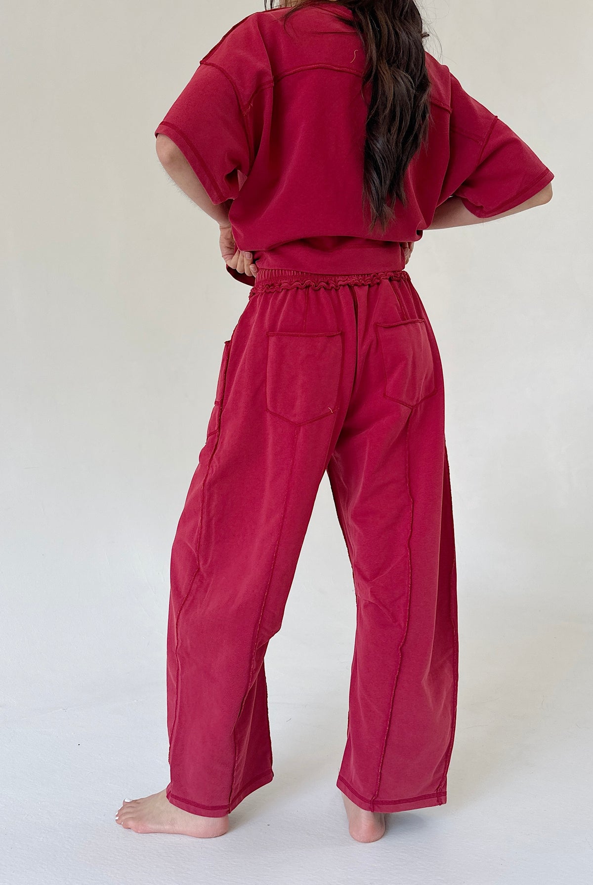 A person shown from the back wearing a matching deep red cropped short-sleeve top and wide-leg pants with large patch pockets, standing barefoot against a plain light background.