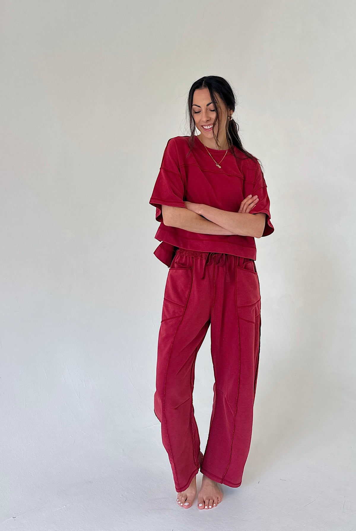 A woman modeling a matching two-piece set in a deep burgundy red, consisting of a short-sleeve top and wide-leg pants, standing against a plain off-white studio background.