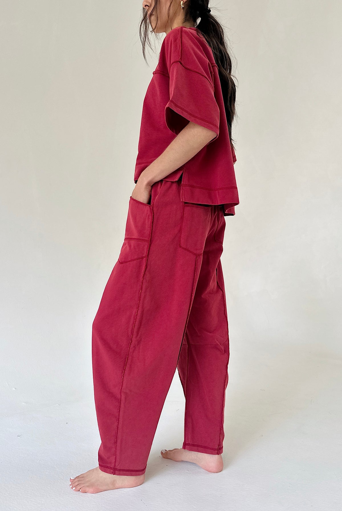 A woman wearing a matching two-piece lounge set in deep magenta, featuring a short-sleeve top and wide-leg pants with side pockets, standing barefoot against a plain off-white background.