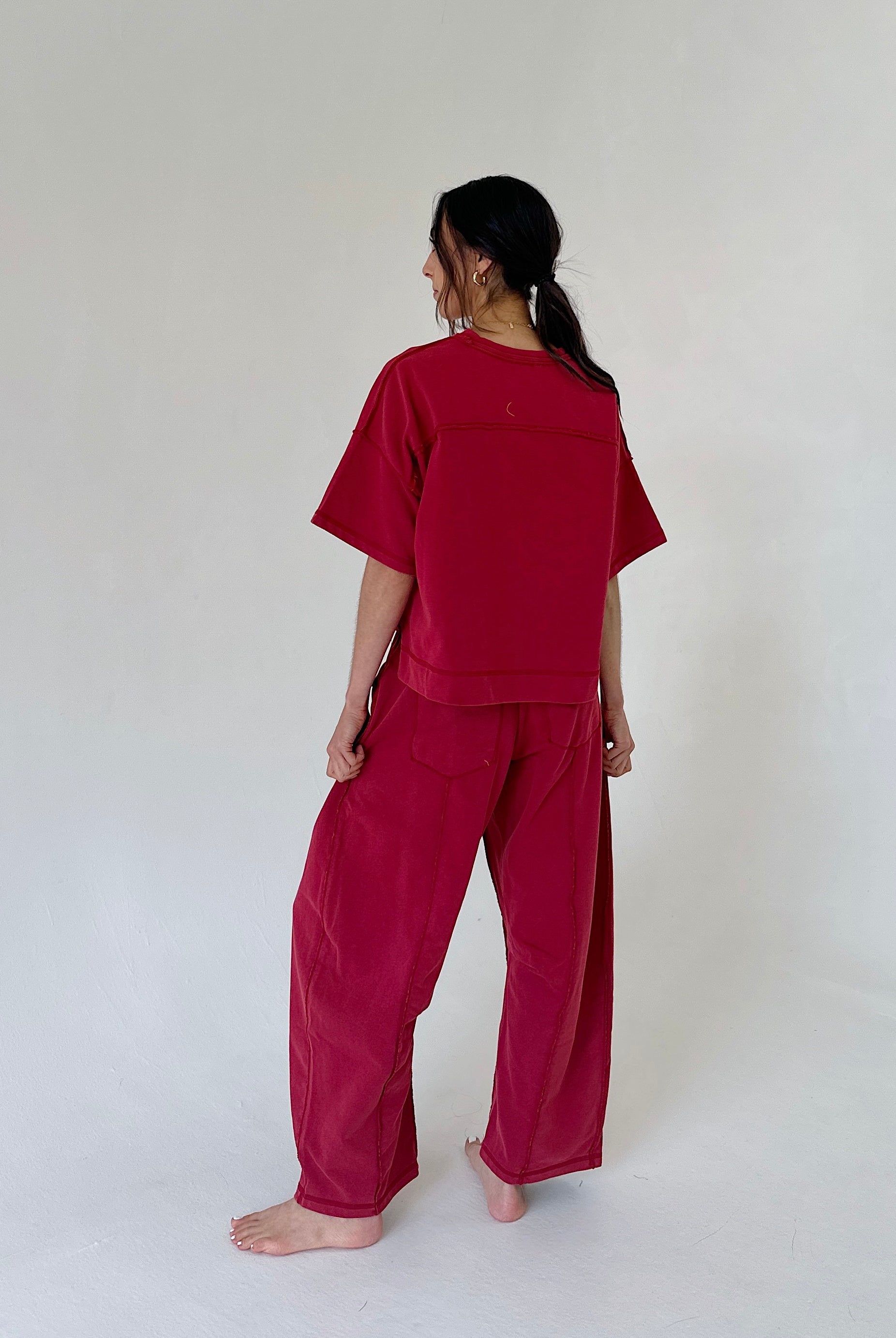 A woman stands barefoot against a plain off-white studio background, viewed from the back, wearing a loose-fitting red short-sleeve top and matching wide-leg pants.