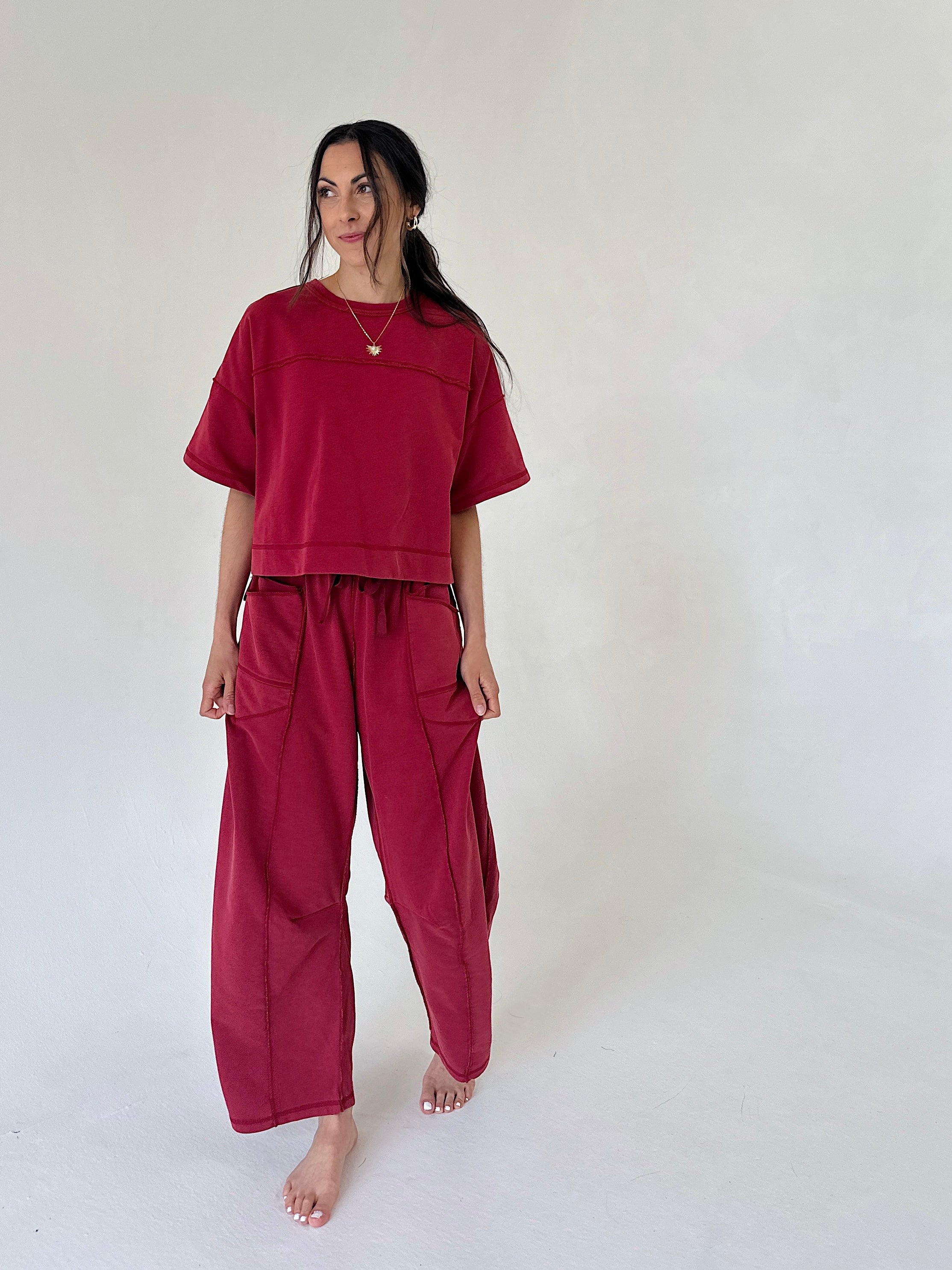 A woman modeling a matching burgundy two-piece lounge set consisting of a short-sleeve cropped top and wide-leg pants, standing against a plain off-white studio background.