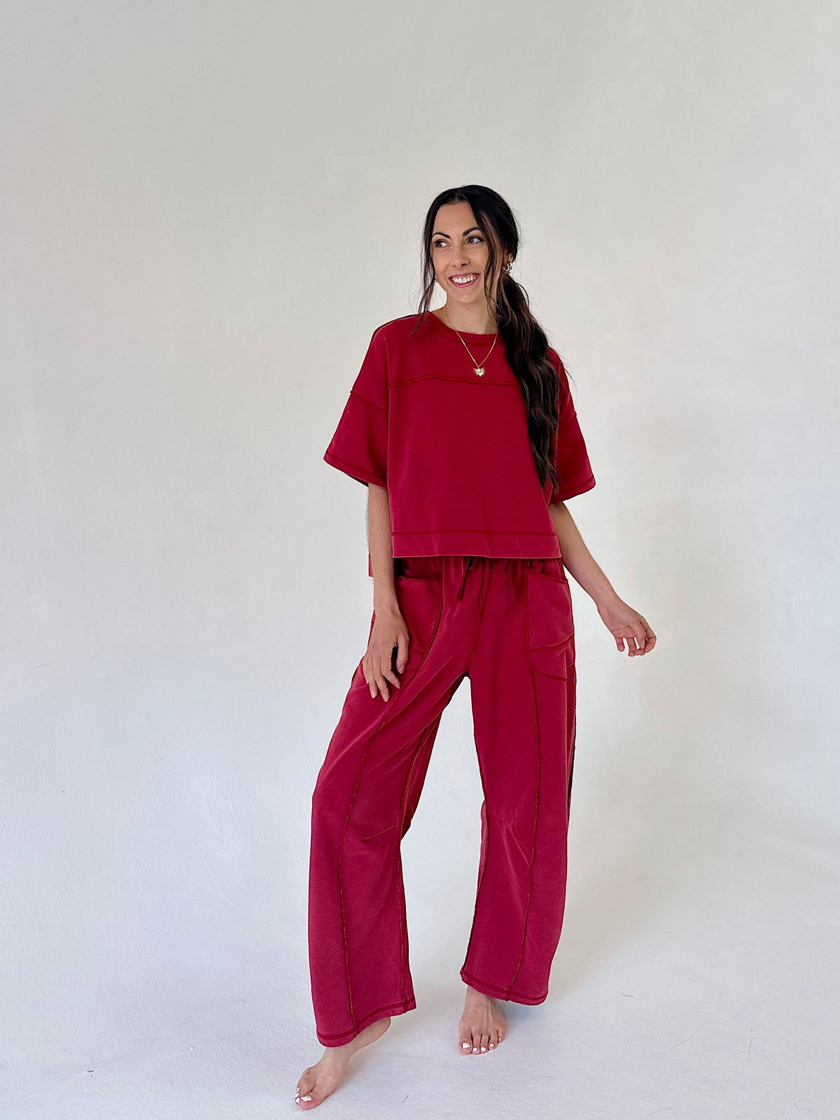 A woman modeling a matching two-piece lounge set in a deep berry red color, consisting of a short-sleeve cropped top and wide-leg pants, standing against a plain off-white studio background.