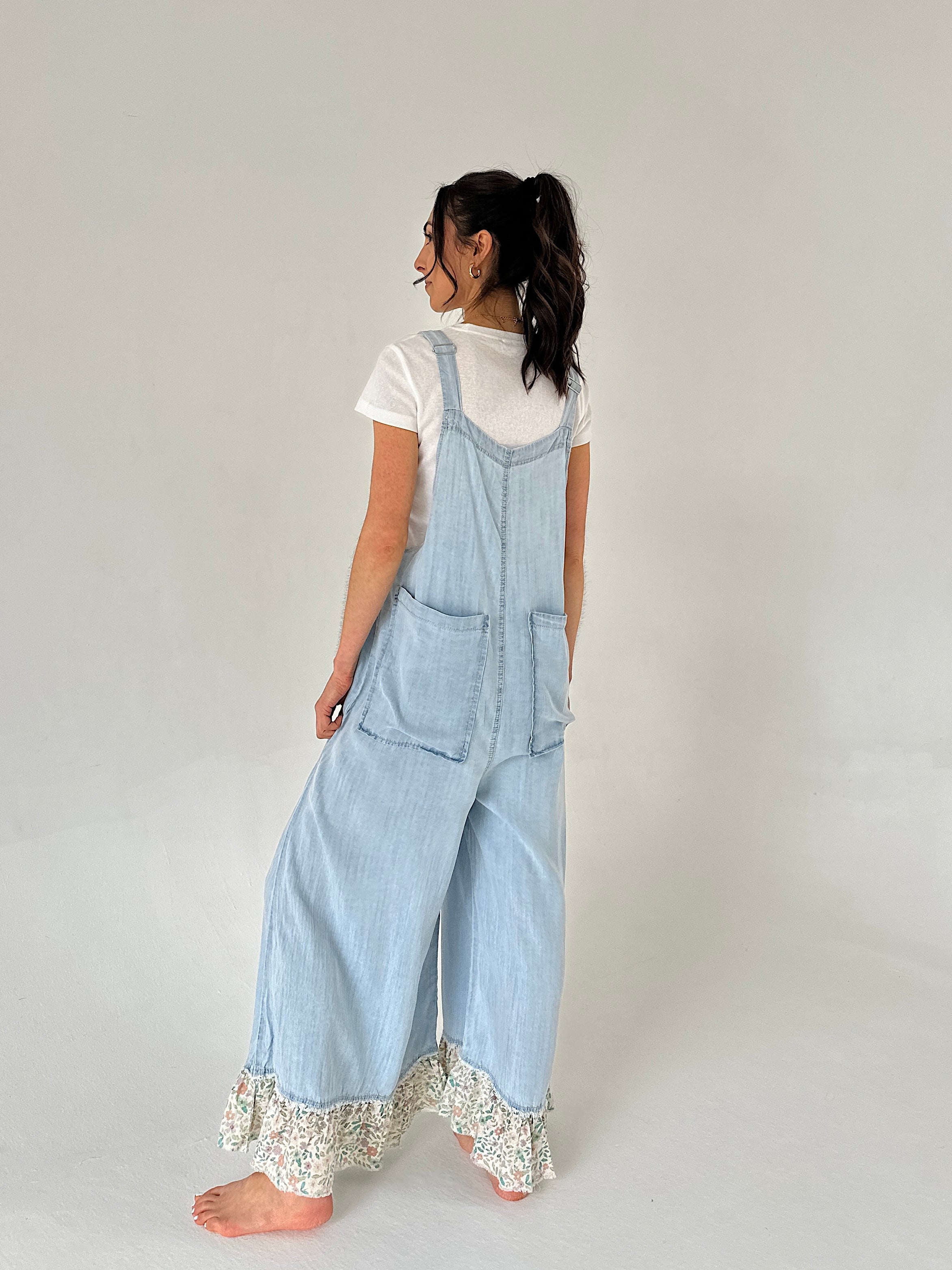 Woman wearing light blue denim overalls with lace trim on a plain background from back