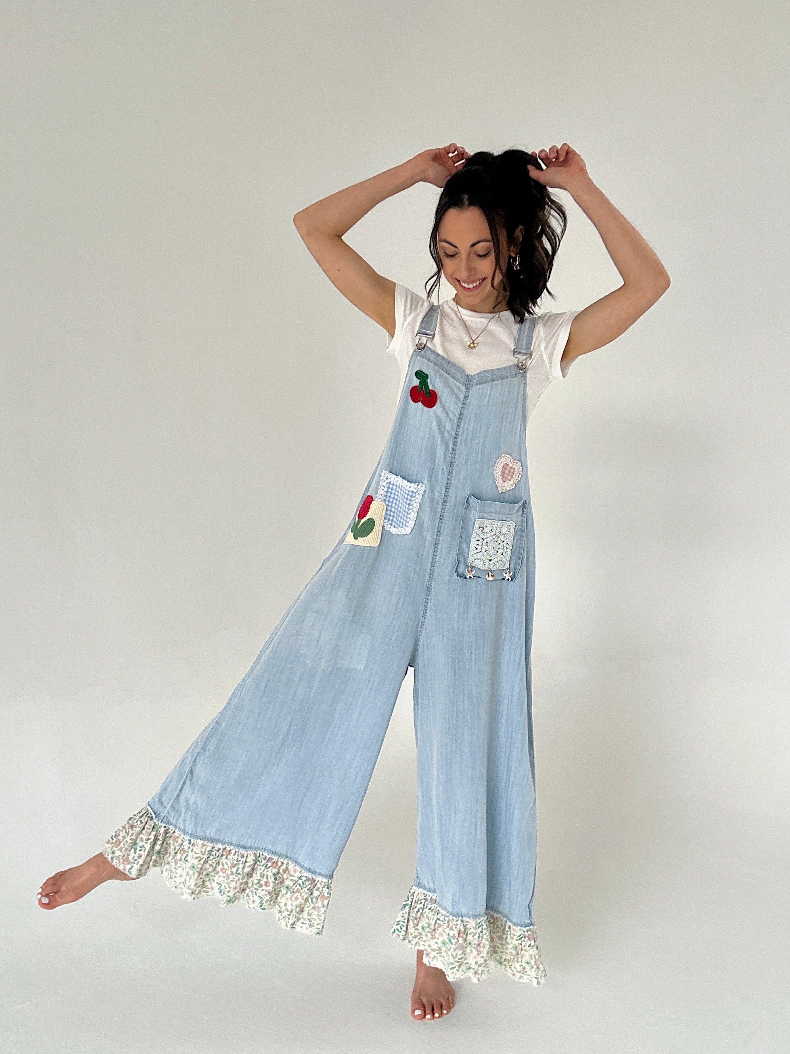 Woman wearing light blue denim overalls with floral trim and embroidered patches on a plain background