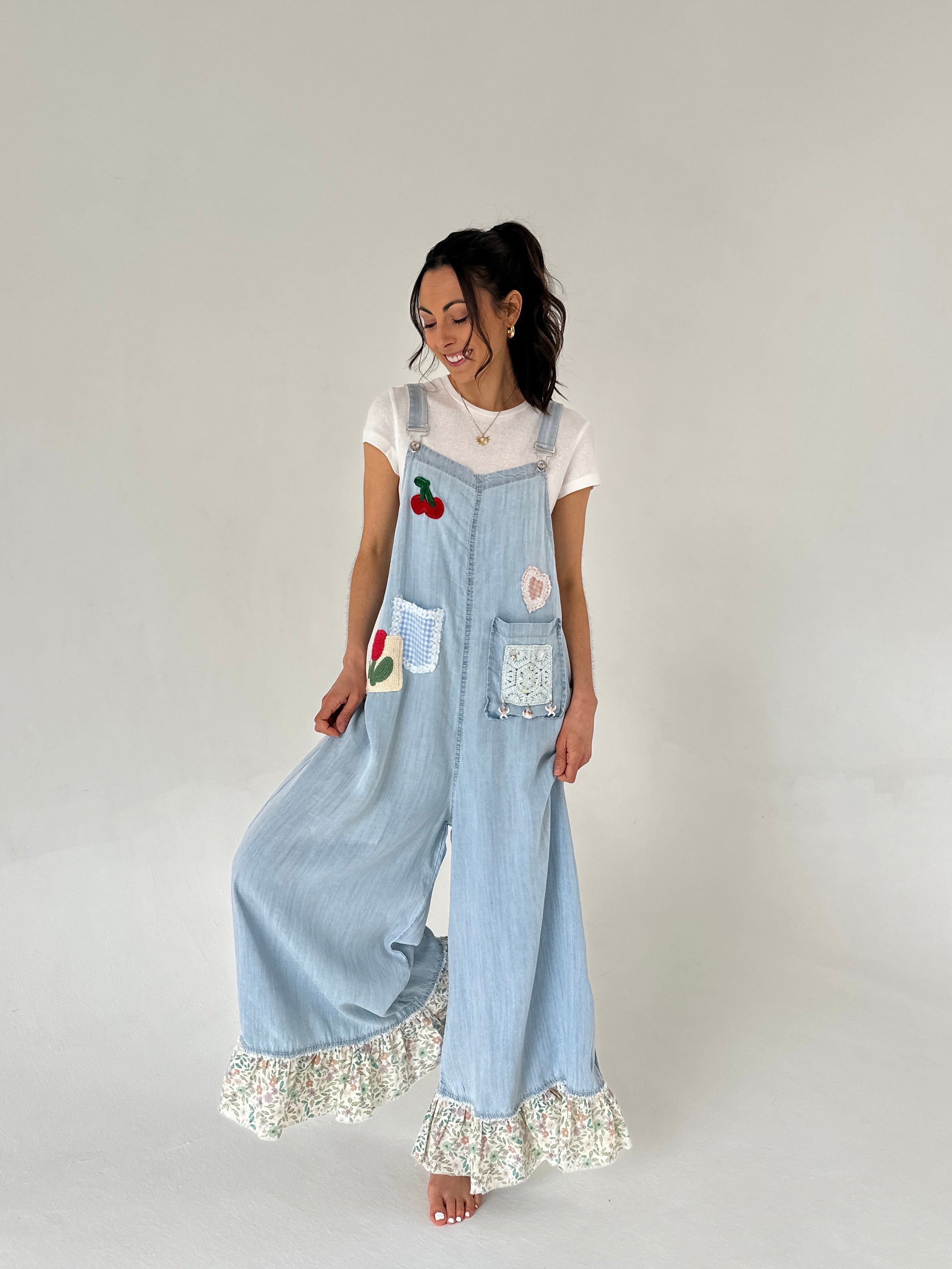 Woman wearing light blue denim overalls with floral hem and patches on a plain background