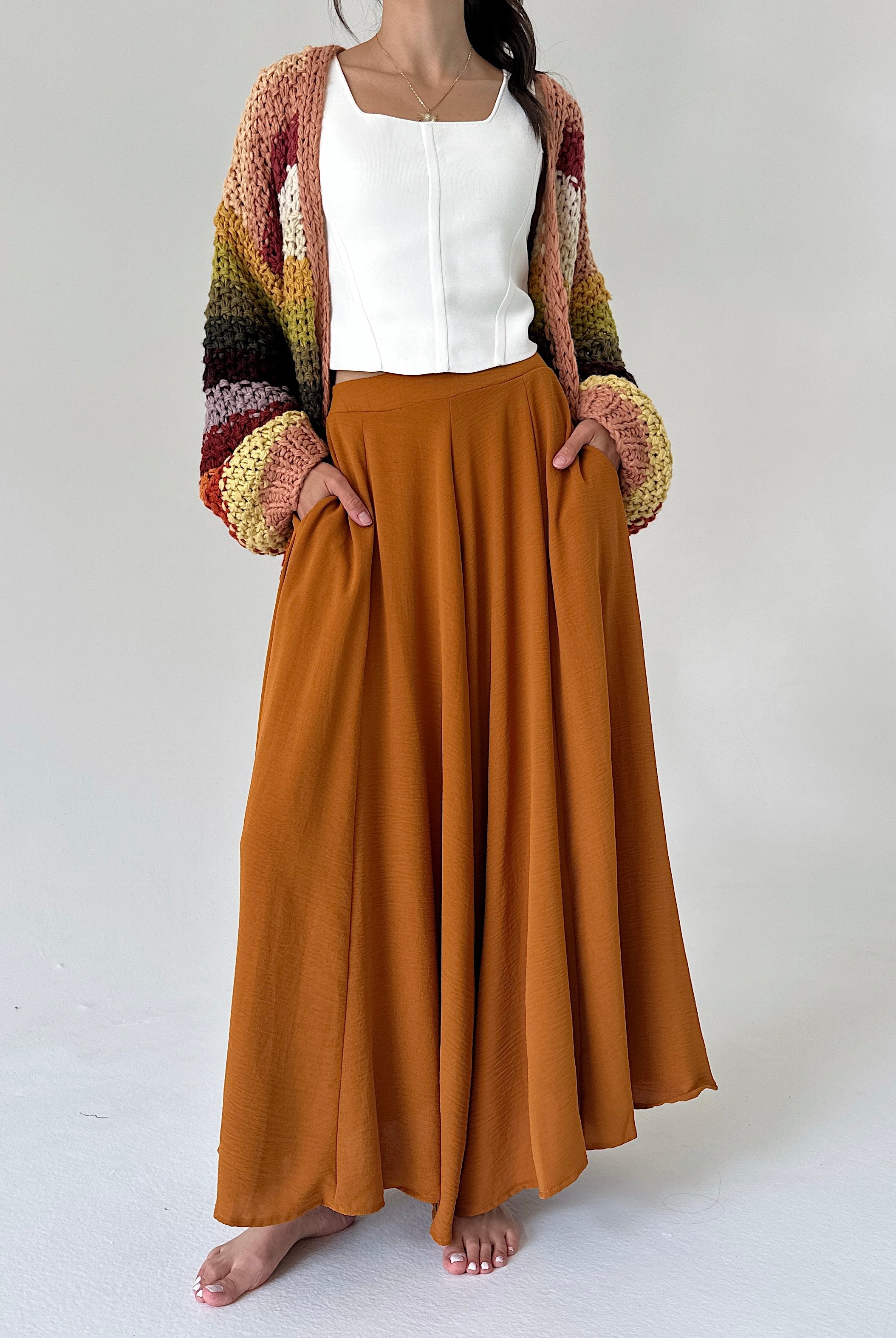 A woman modeling a long, flowing rust-orange wide leg pants with side pockets, paired with a white sleeveless top and a chunky multicolor knit cardigan with striped sleeves, standing against a light gray studio background.