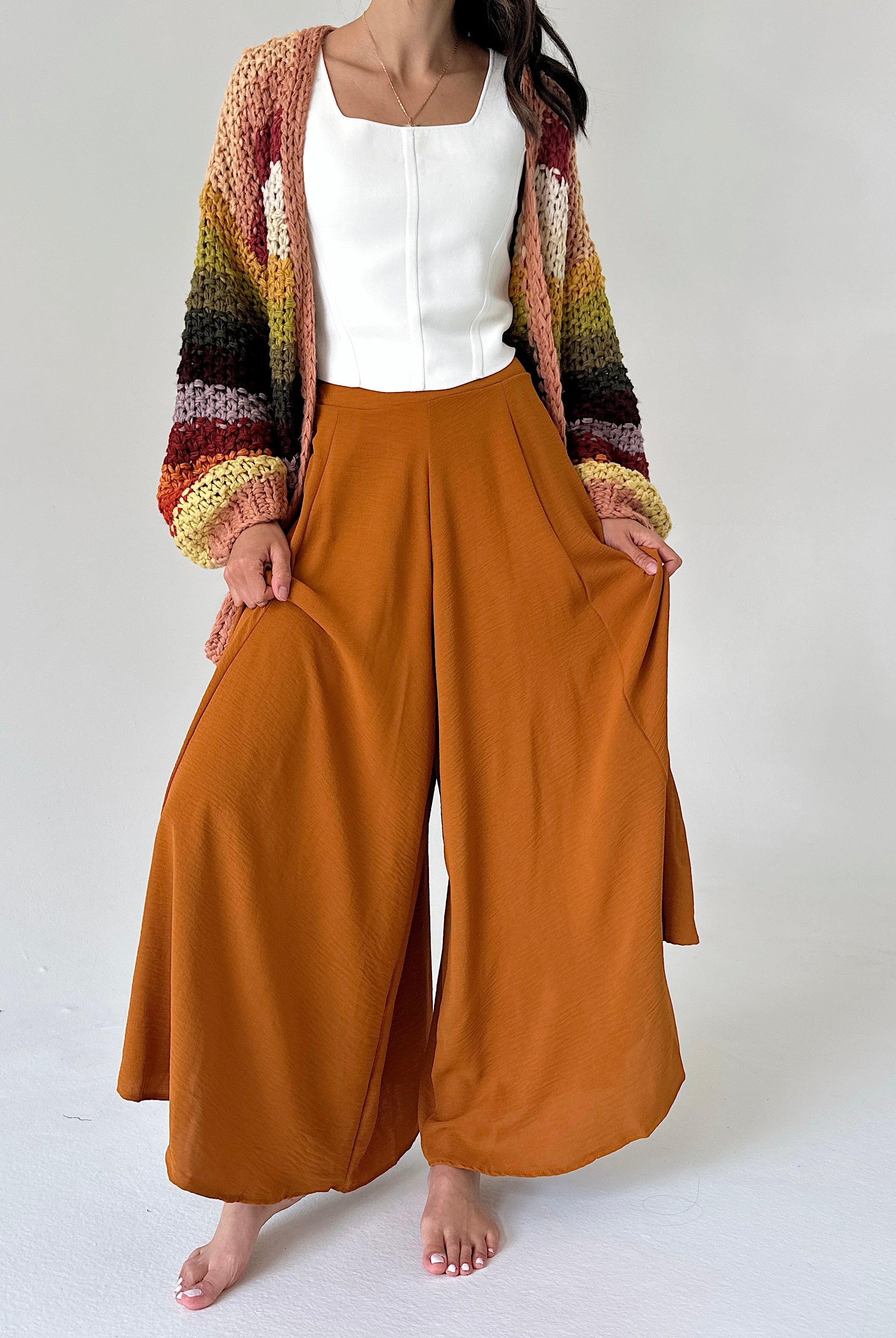 A woman modeling a pair of wide-leg rust-colored pants with a white top and a multicolor striped knit cardigan, standing against a plain light gray background.