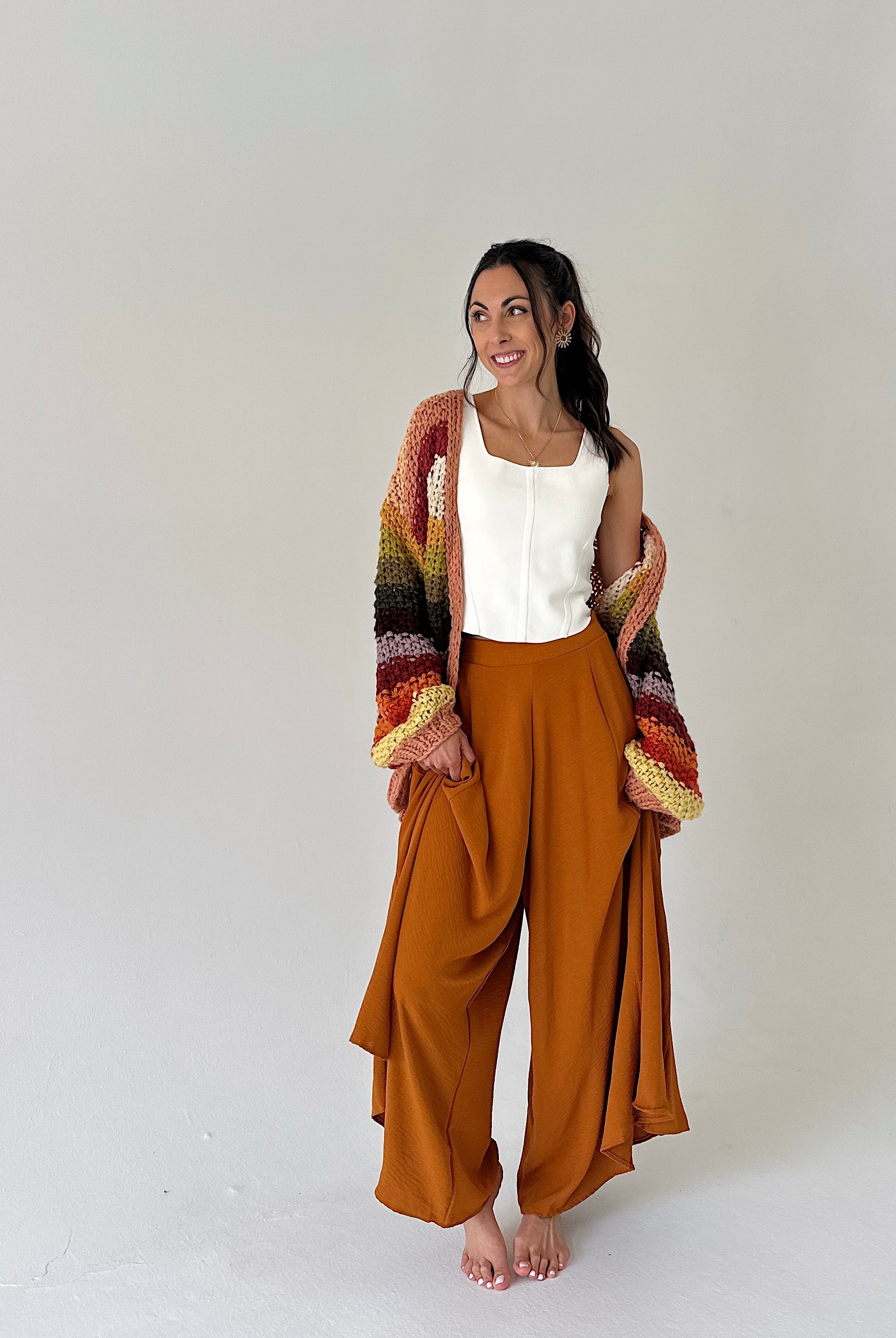 A woman stands barefoot against a plain light gray studio background, wearing a white sleeveless top, wide-leg rust-brown pants, and a chunky multicolor knit cardigan draped off her shoulders.