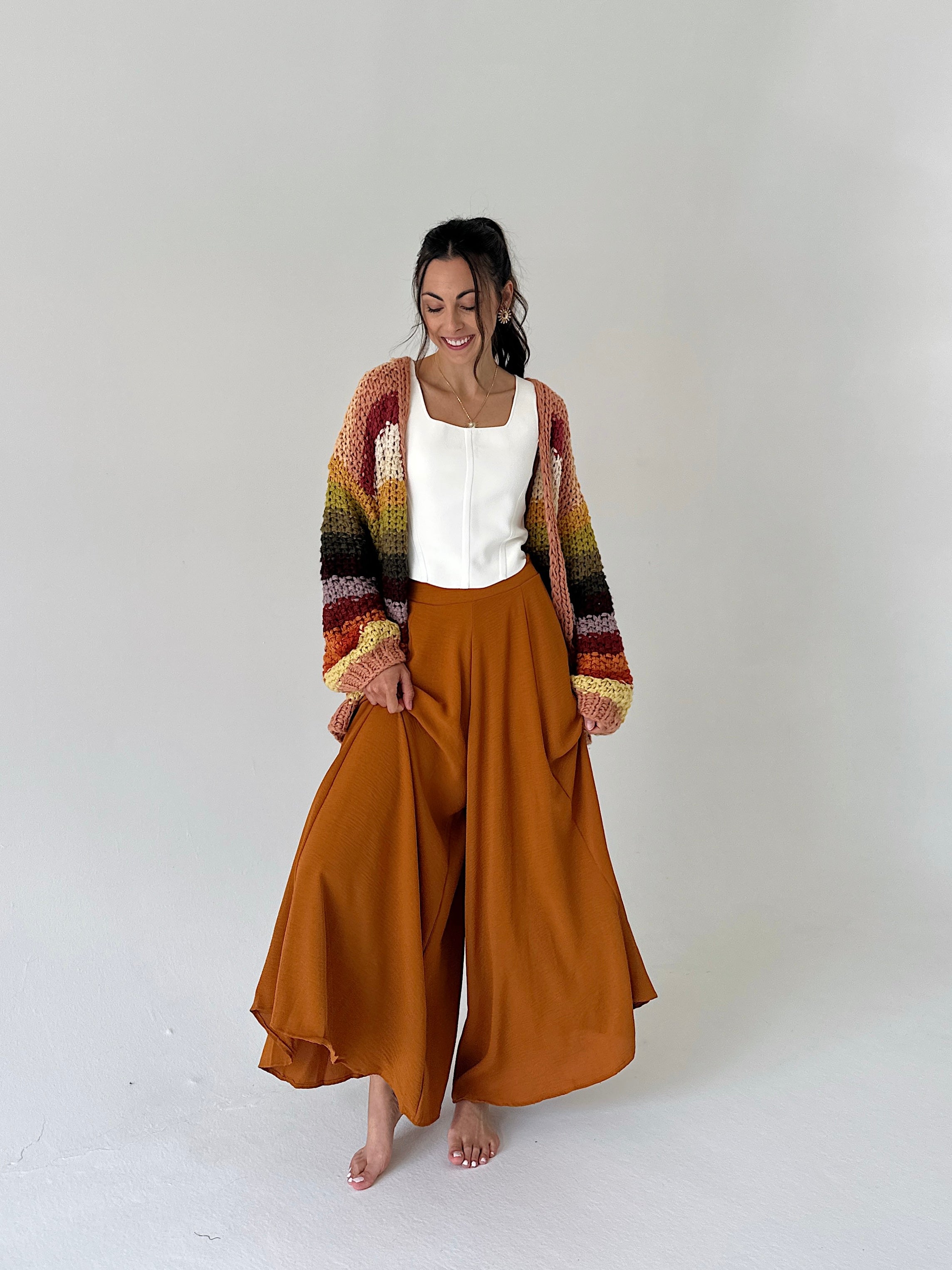 A woman stands barefoot against a plain light gray studio background, wearing a cream sleeveless top, wide-leg rust-orange pants, and a multicolored striped knit cardigan.