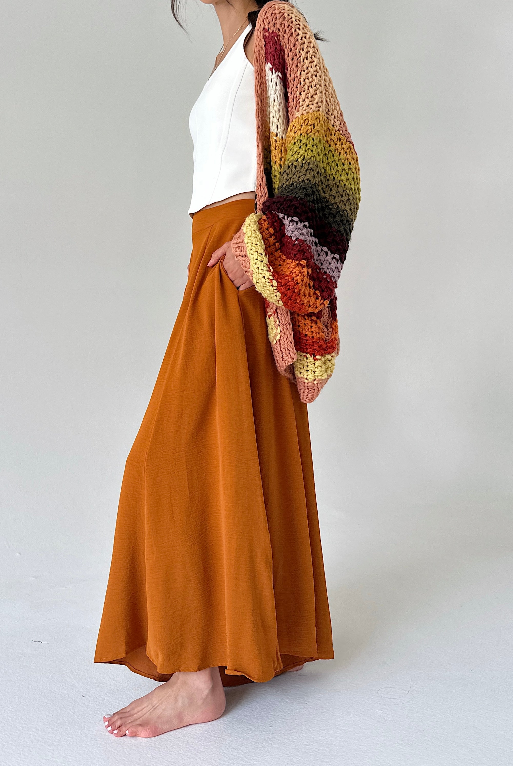 A model wearing a rust-orange maxi pant, a white top, and a chunky multicolor crochet cardigan draped over one shoulder, standing barefoot against a light gray studio background.