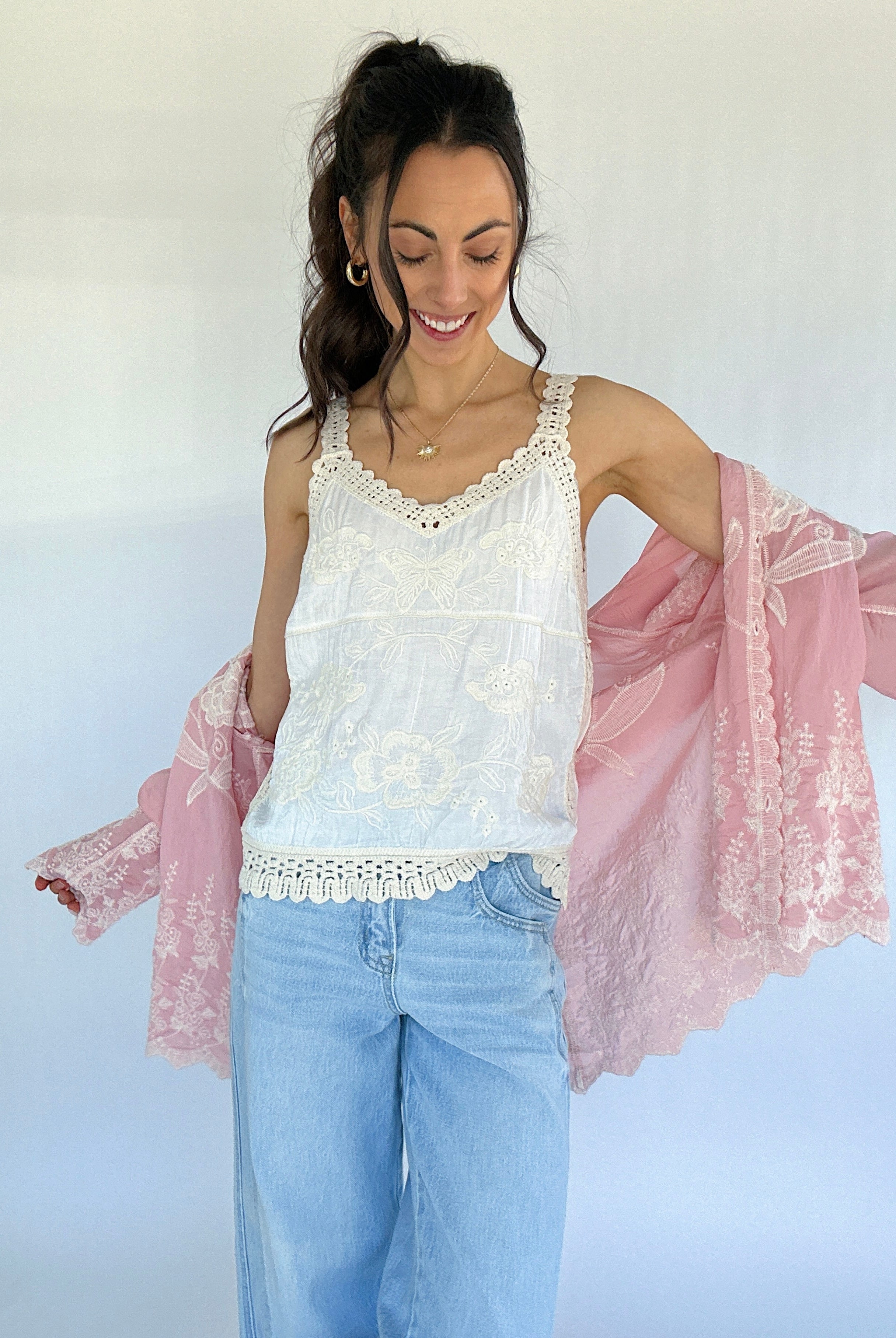 Woman wearing a white lace top and light blue jeans with a pink lace long sleeve over her shoulders against a white background.