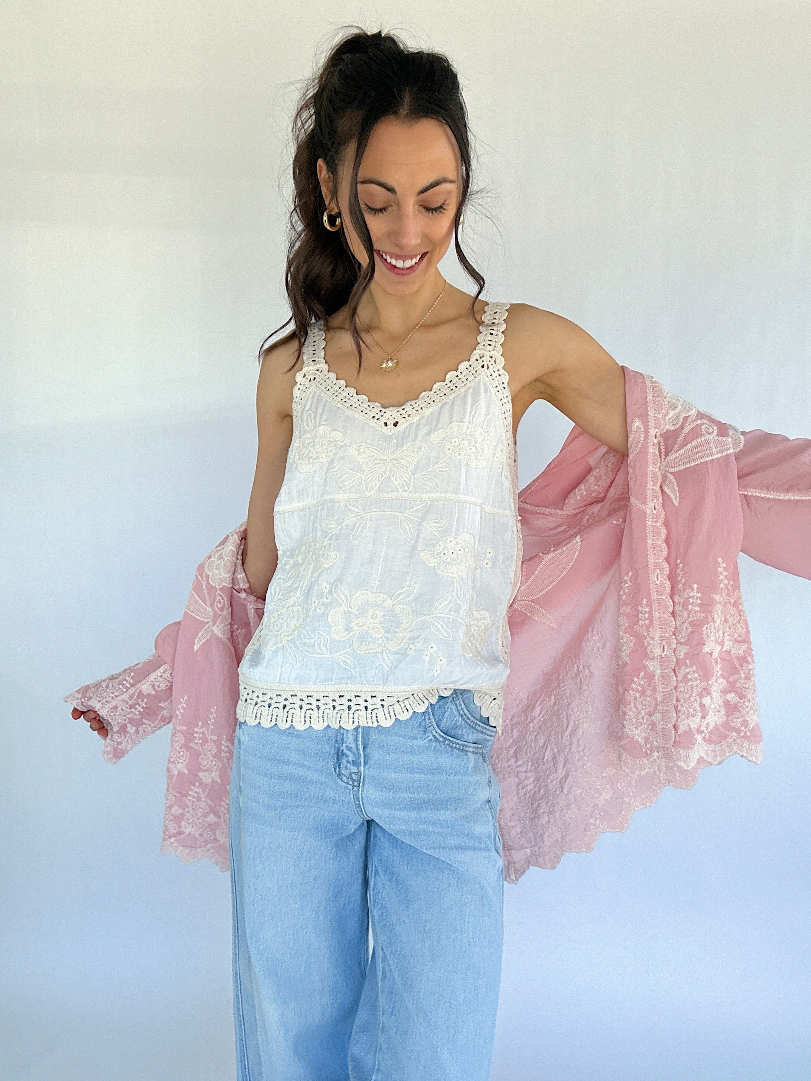 Woman wearing a white lace top and light blue jeans with a pink lace long sleeve over her shoulders against a white background.