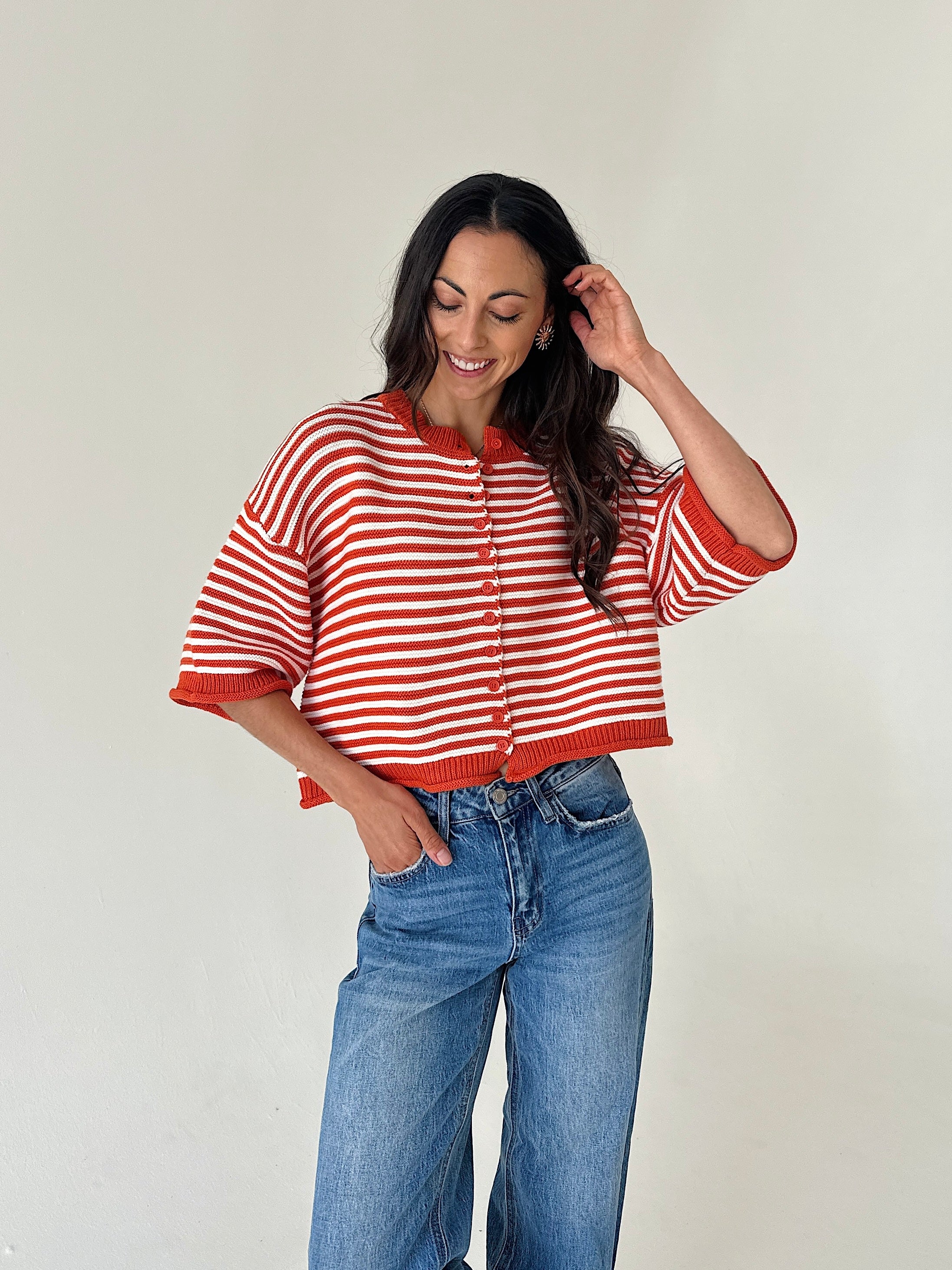 A woman modeling a cropped orange and white striped knit cardigan with blue jeans, standing against a plain off-white wall.