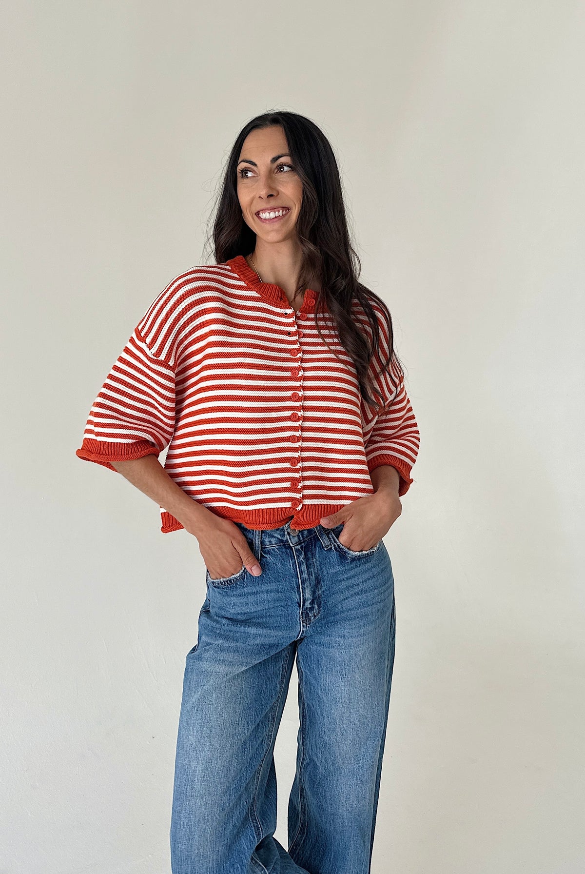 A woman modeling an orange and white striped knit cardigan with a drawstring neckline, paired with blue jeans, against a plain off-white wall.