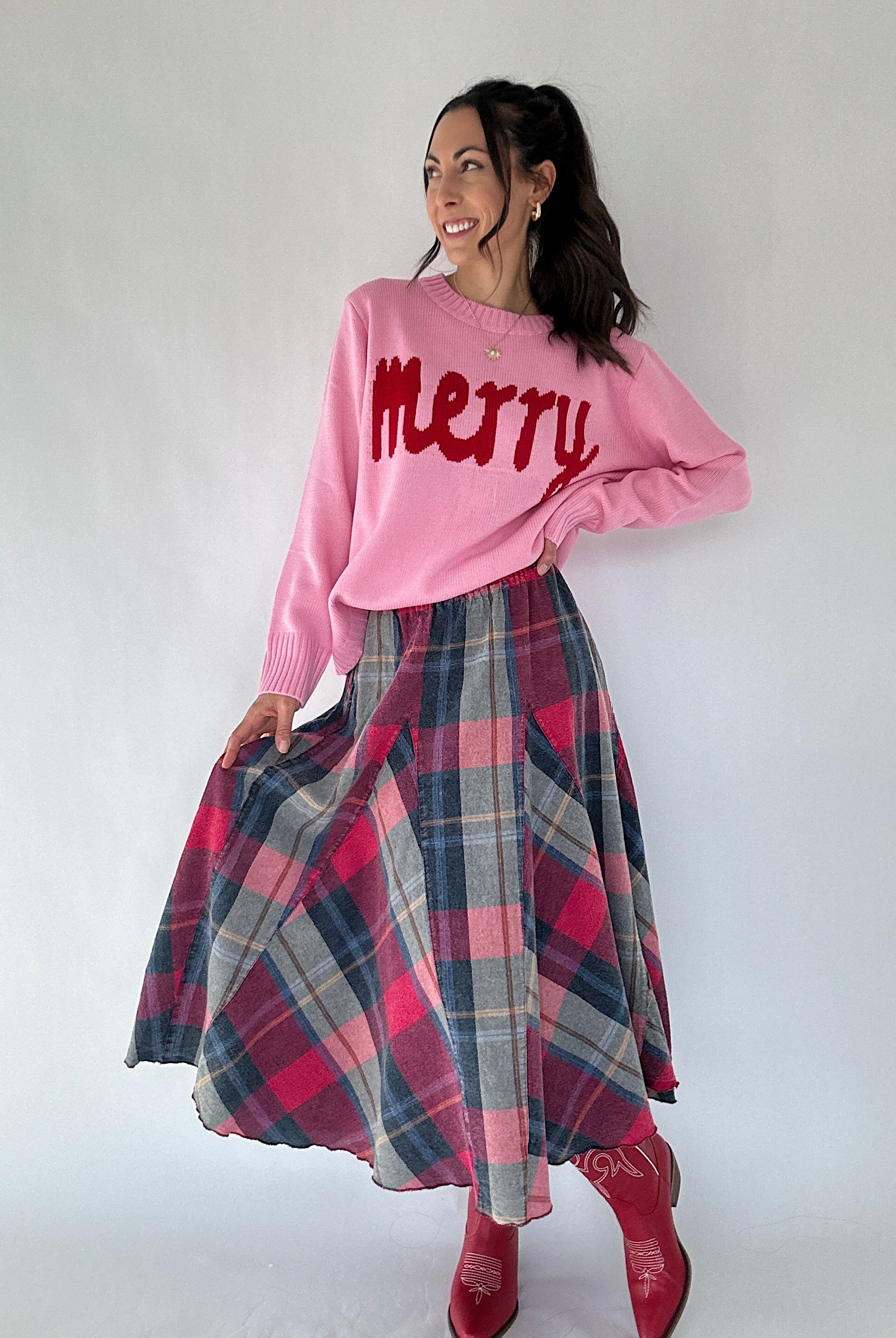 Woman wearing a pink 'Merry' sweater and plaid skirt on a white background