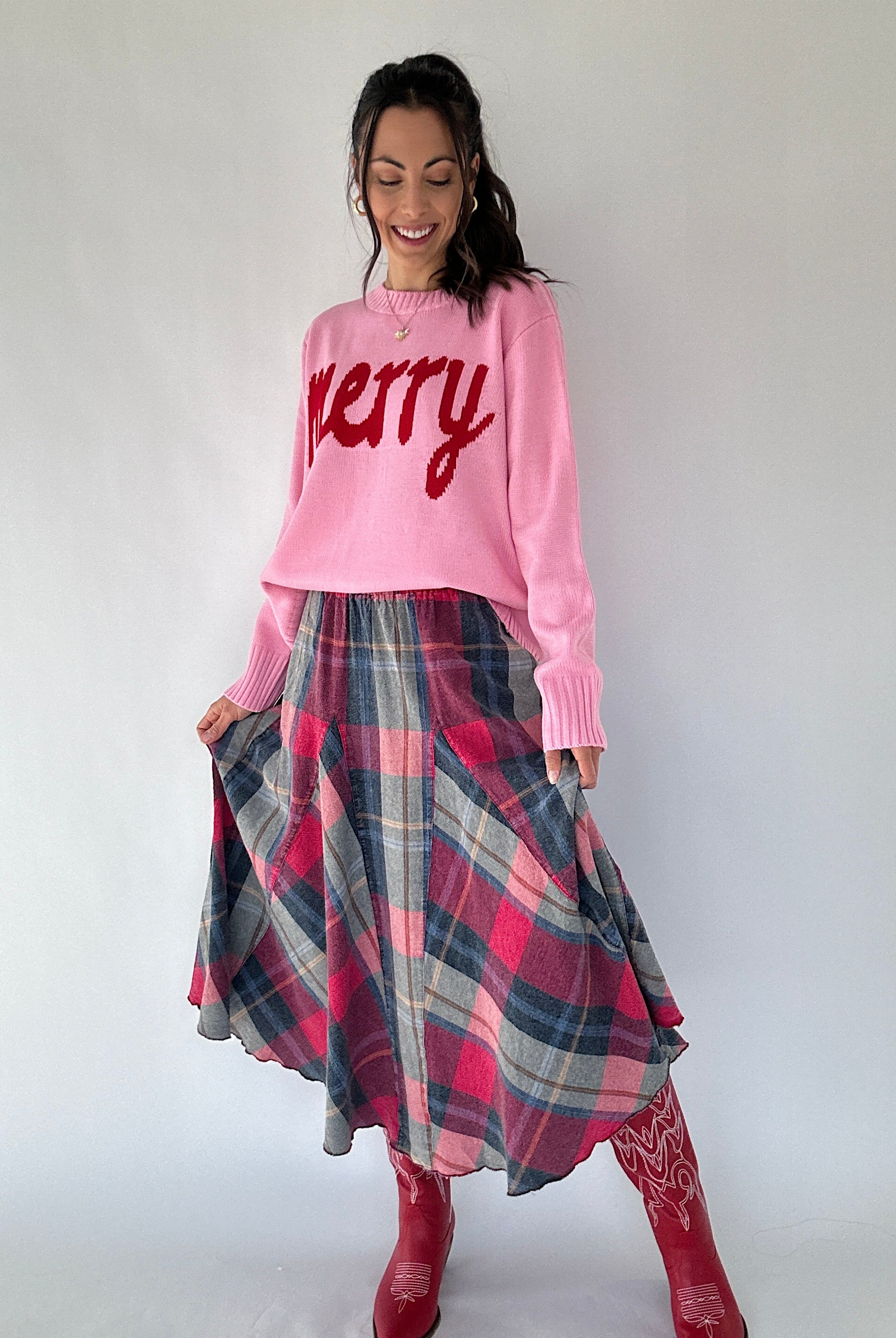 Woman wearing a pink sweater with 'Merry' text and a plaid skirt on a white background