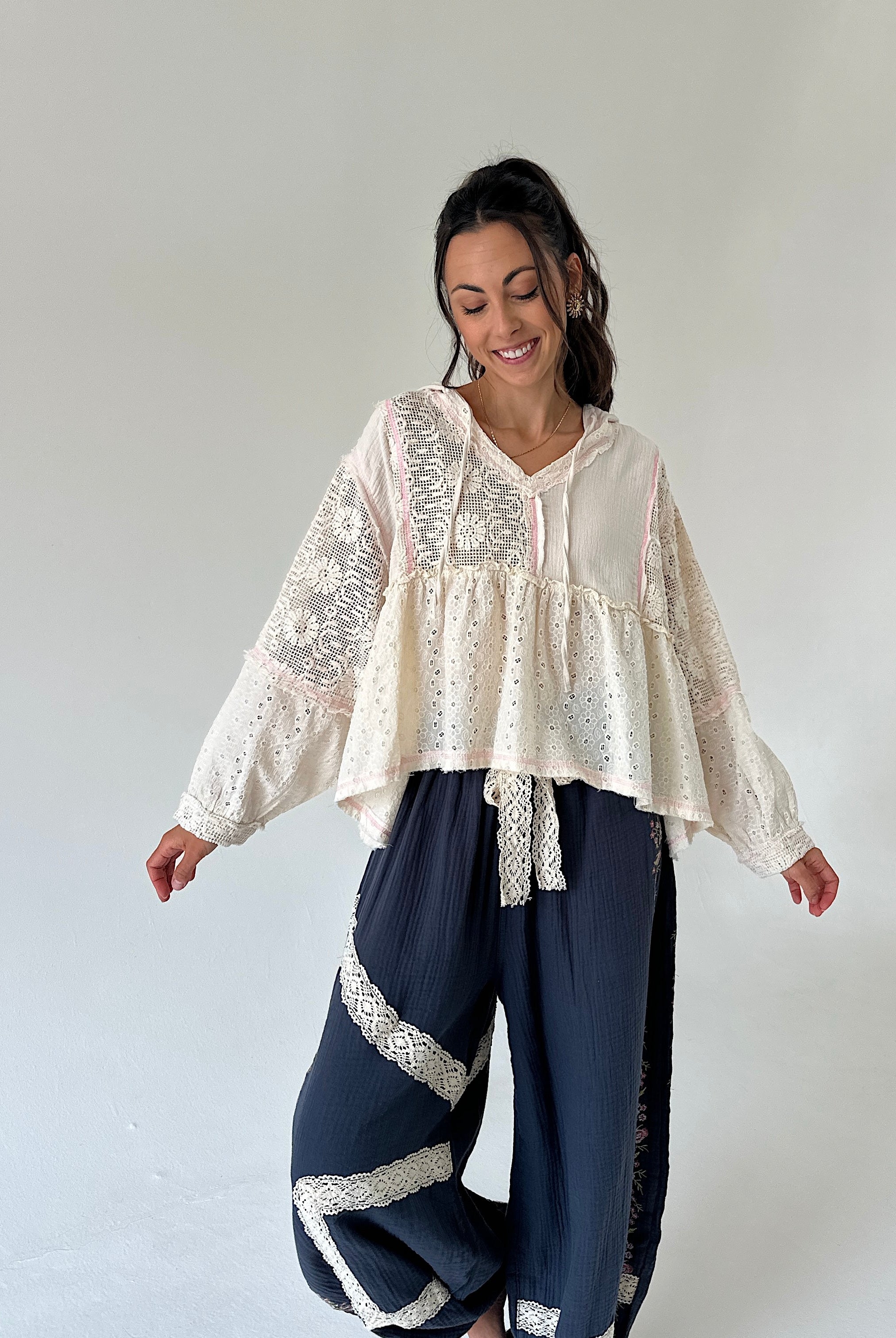 A woman modeling a bohemian-style outfit consisting of a cream lace and dot-print long-sleeve top paired with navy blue wide-leg pants featuring white lace trim, set against a plain light gray studio background.