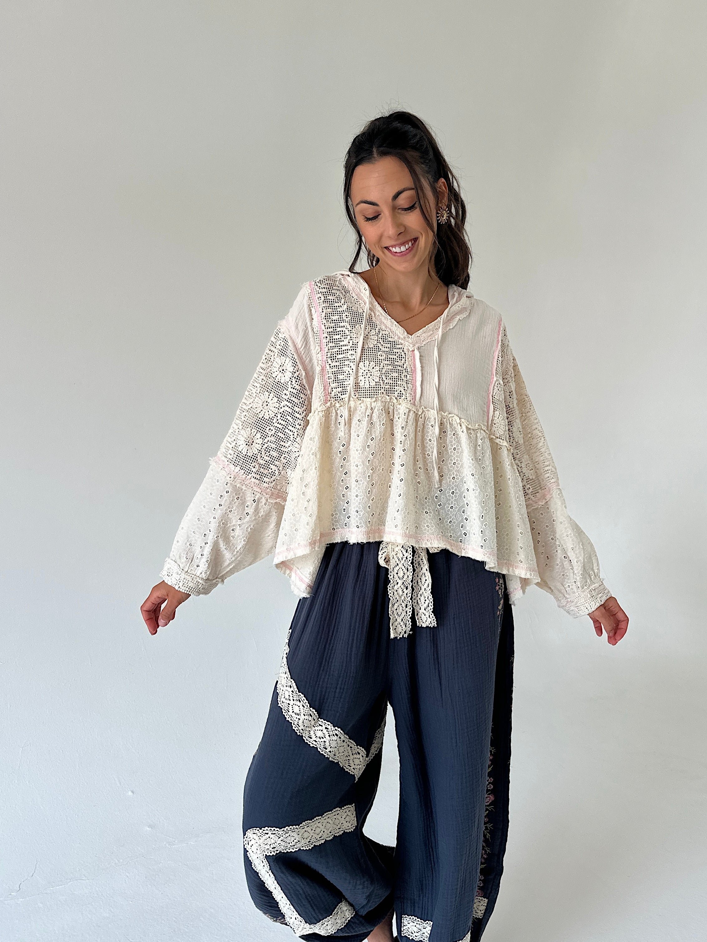 A woman modeling a bohemian-style outfit consisting of a cream lace and dot-print long-sleeve top paired with navy blue wide-leg pants featuring white lace trim, set against a plain light gray studio background.