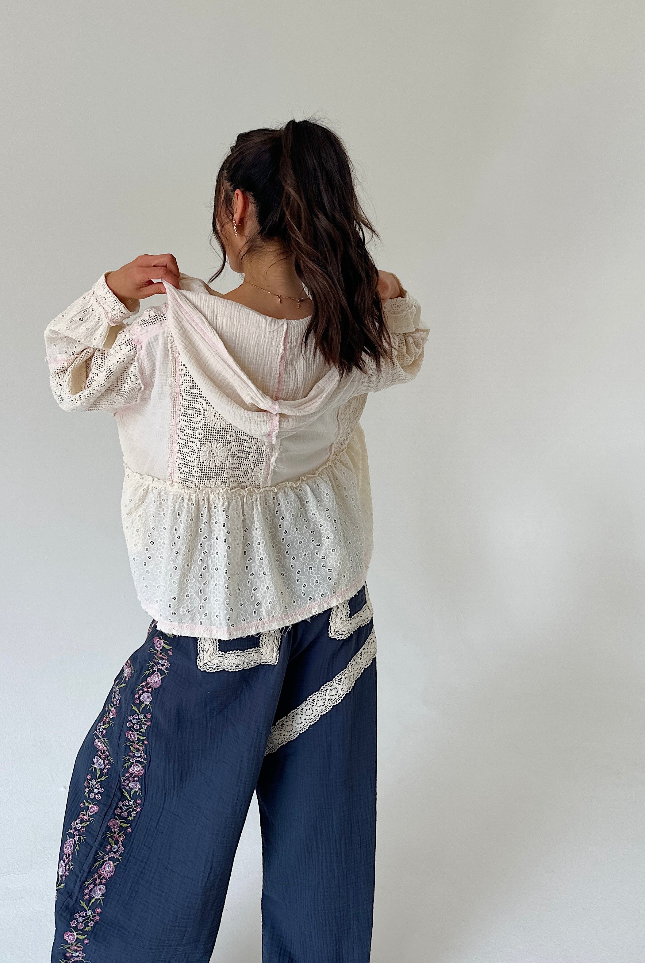 A woman shown from behind wearing a cream eyelet lace long-sleeve top and dark blue wide-leg pants with floral embroidery, posing against a plain light gray wall.