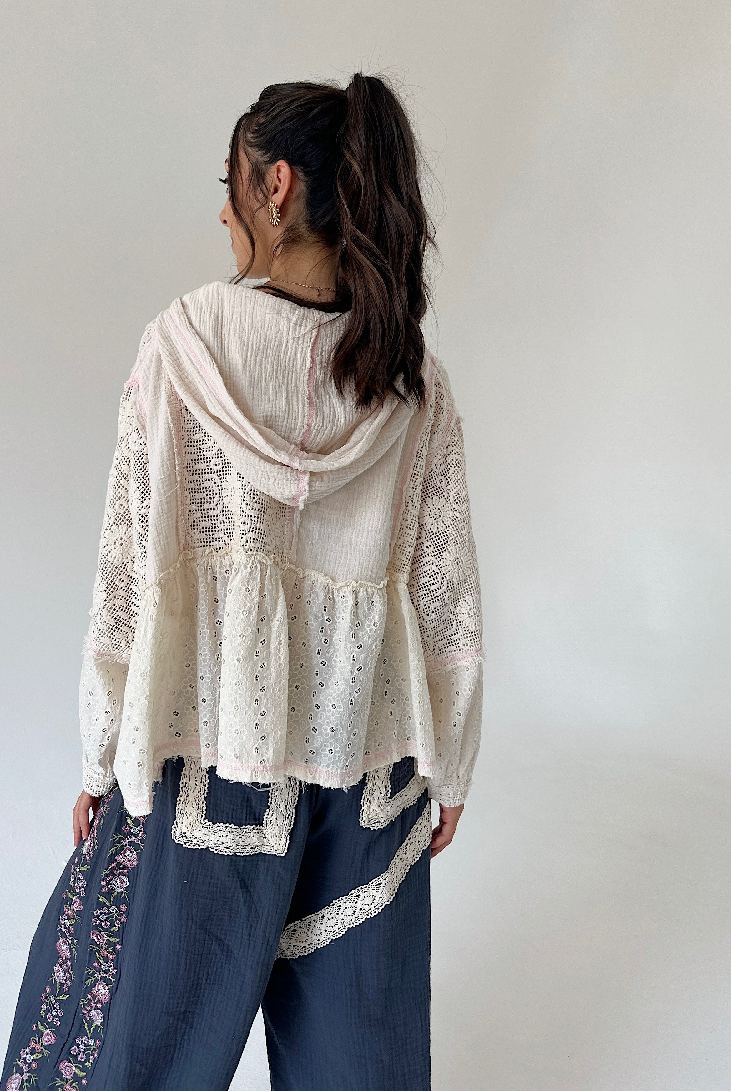 A woman shown from the back wearing a cream-colored lace and eyelet hooded top layered over dark blue pants with floral embroidery and lace trim, set against a plain light gray background.