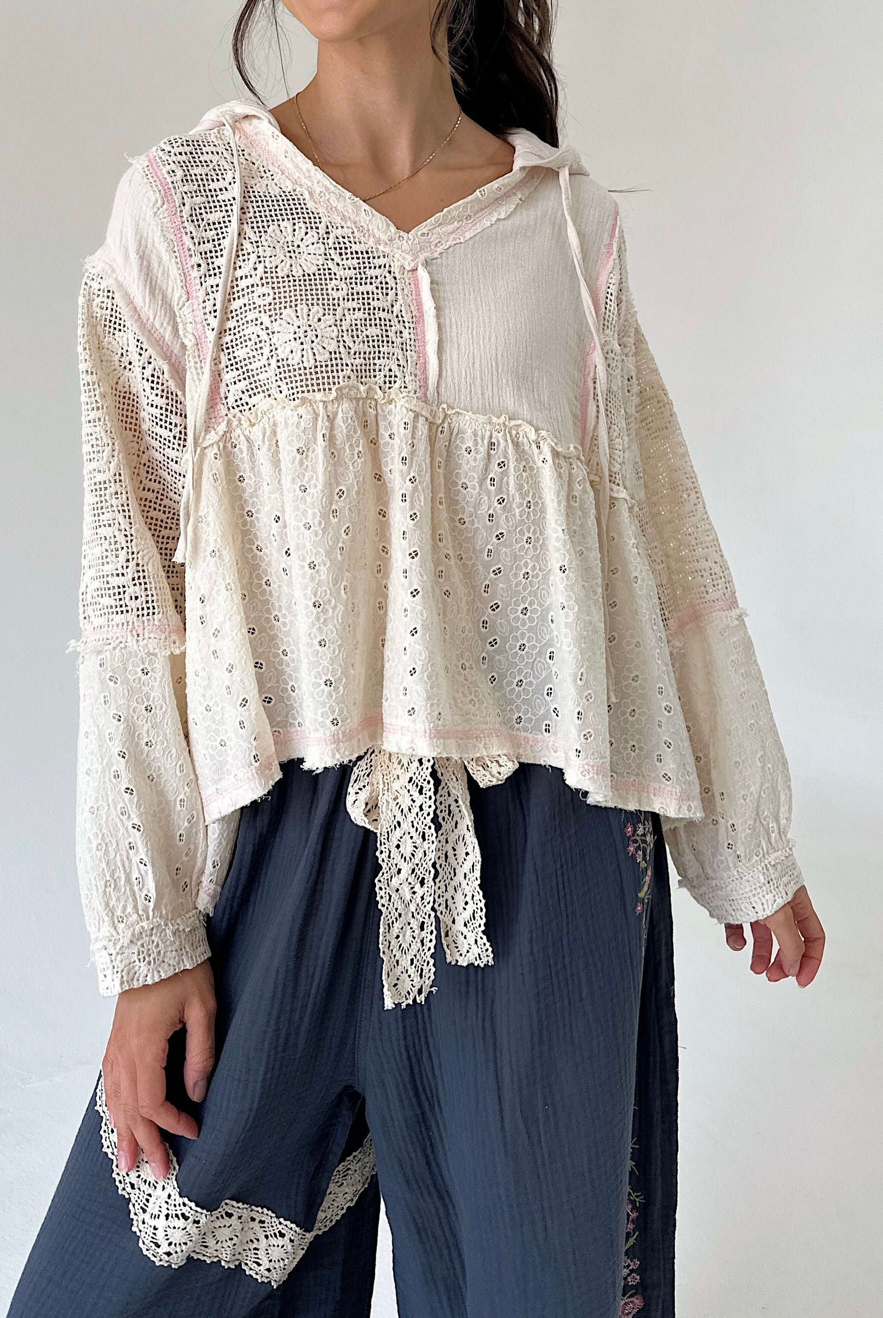 A woman wearing a cream eyelet lace long-sleeve blouse with a tie waist and dark blue wide-leg pants against a plain white background.